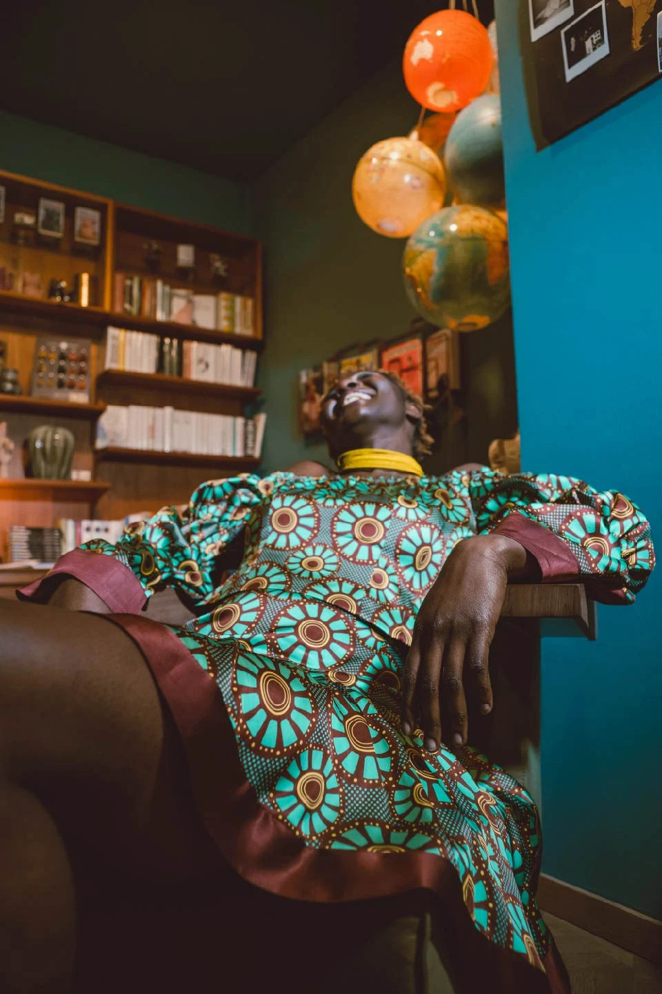 una mujer riéndose, recostada sobre una silla, lleva puesto un vestido estampado con un diseño de estilo africano en colores bordeaux, turquesa y amarillo
