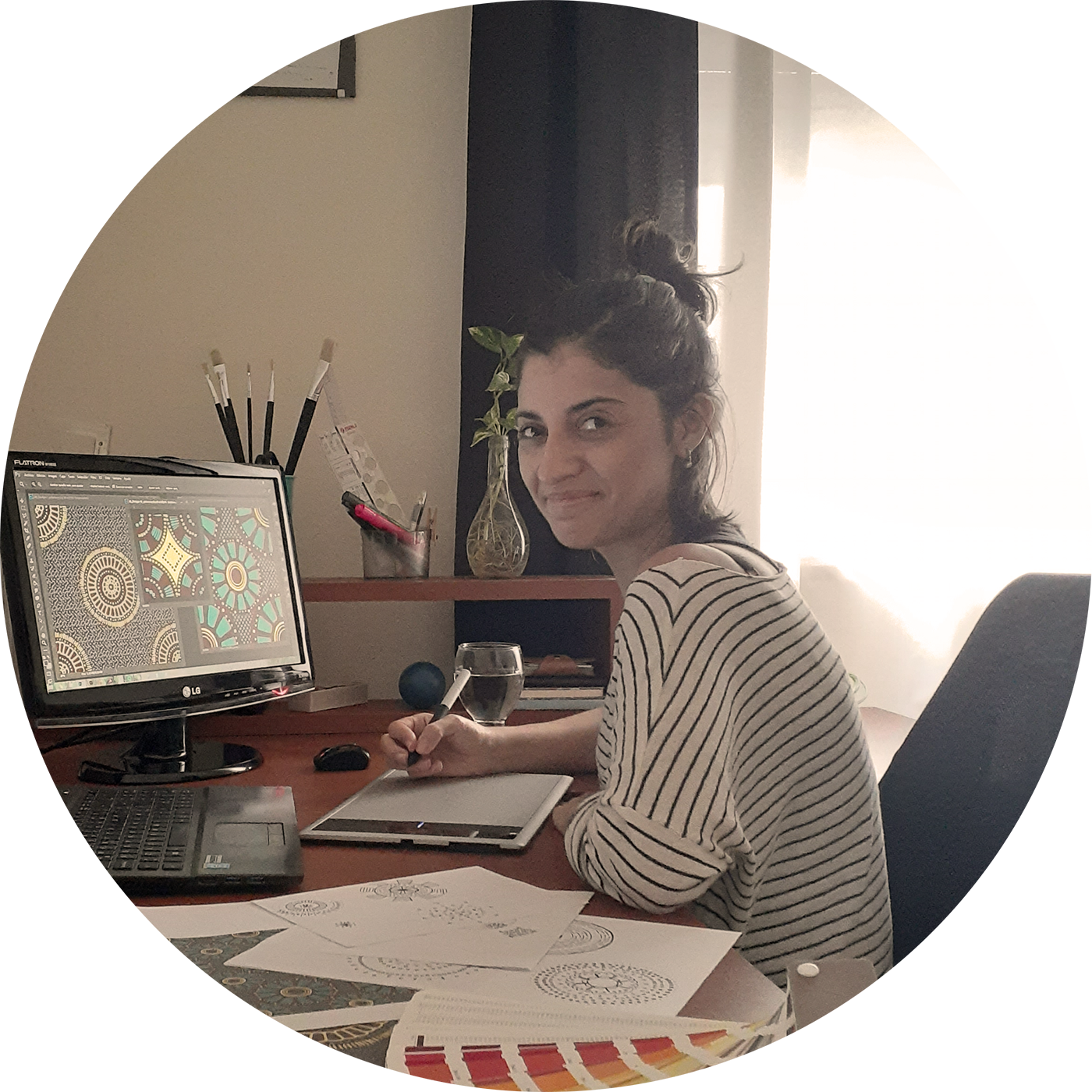 una diseñadora textil mira a la cámara sonriendo, sentada en su escritorio de trabajo frente a un monitor con diseños, usando una tableta gráfica digitalizadora, con bocetos, dibujos y cartas de color sobre la mesa