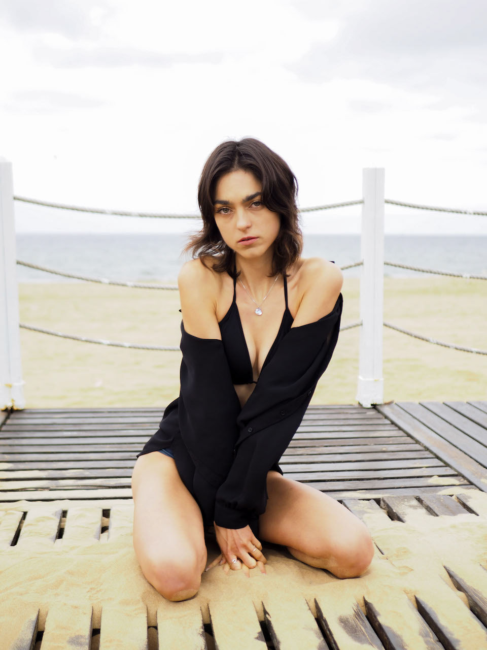 A woman sits on a sandy beach boardwalk with a black shirt draped over her shoulders, gazing intently at the camera against a backdrop of calm seas and overcast skies