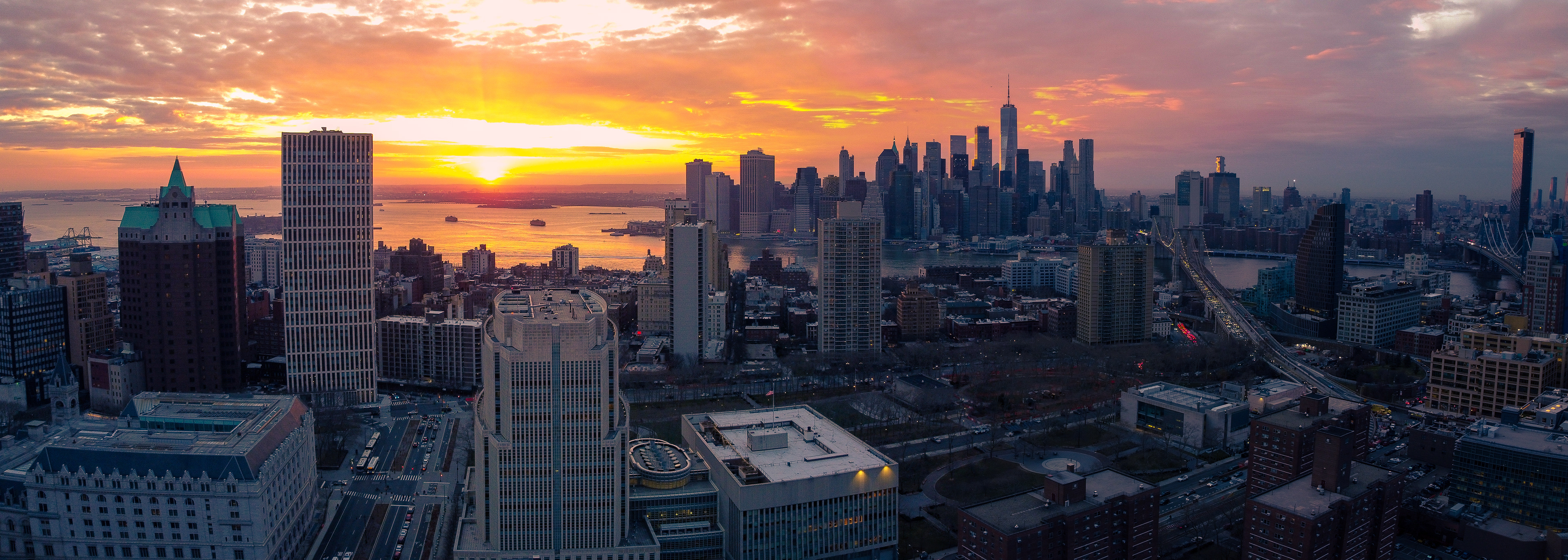 A north-western facing panoramic aerial view of  downtown Brooklyn, New  York with the Manhattan skyline behind it and a glowing orange and pink sunset behind that. 