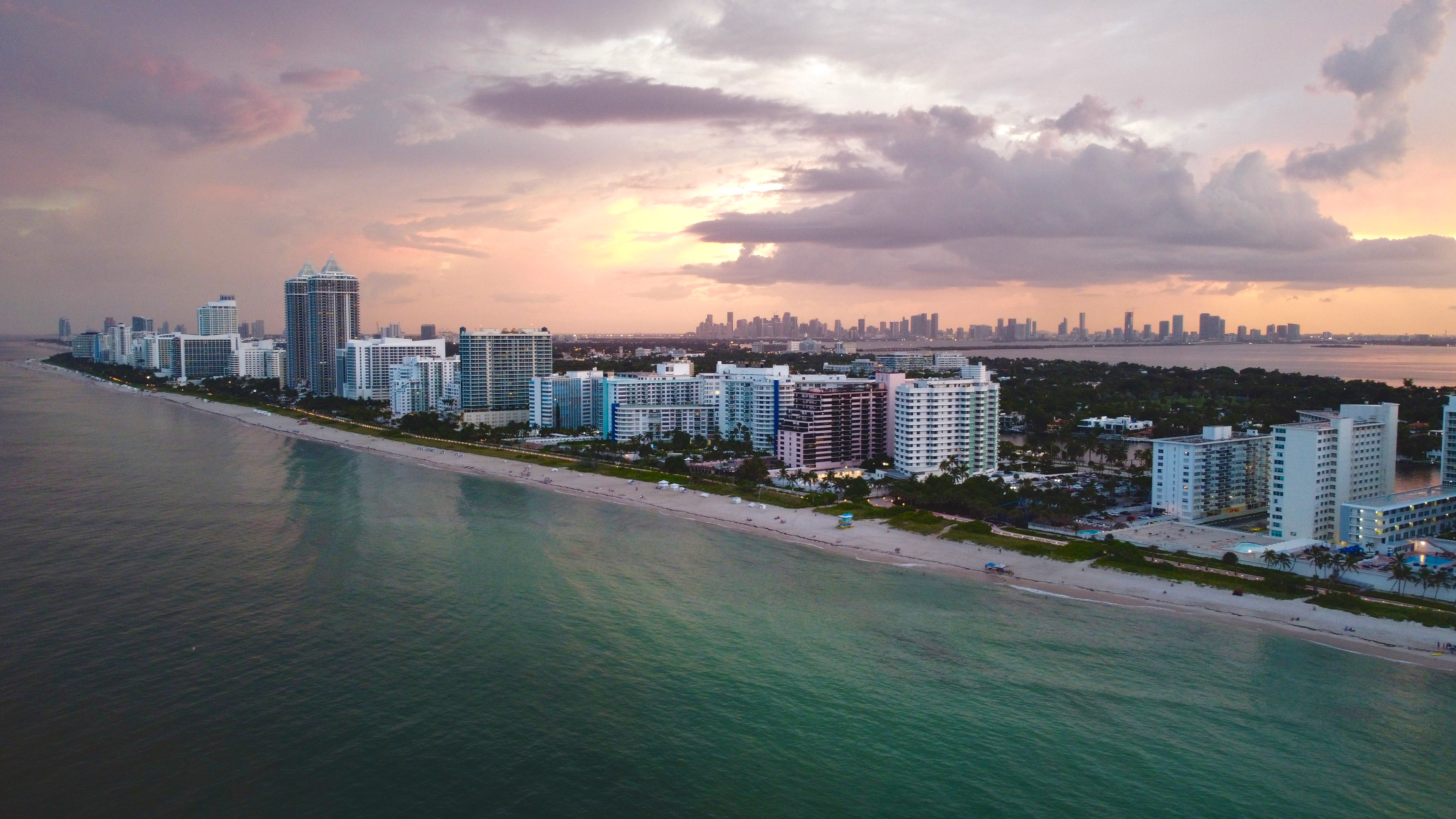 A panoramic aerial view of a sunset behind Miami Beach, Florida. The waters are teal and rippled. The sky is cotton candy pink and purple. 