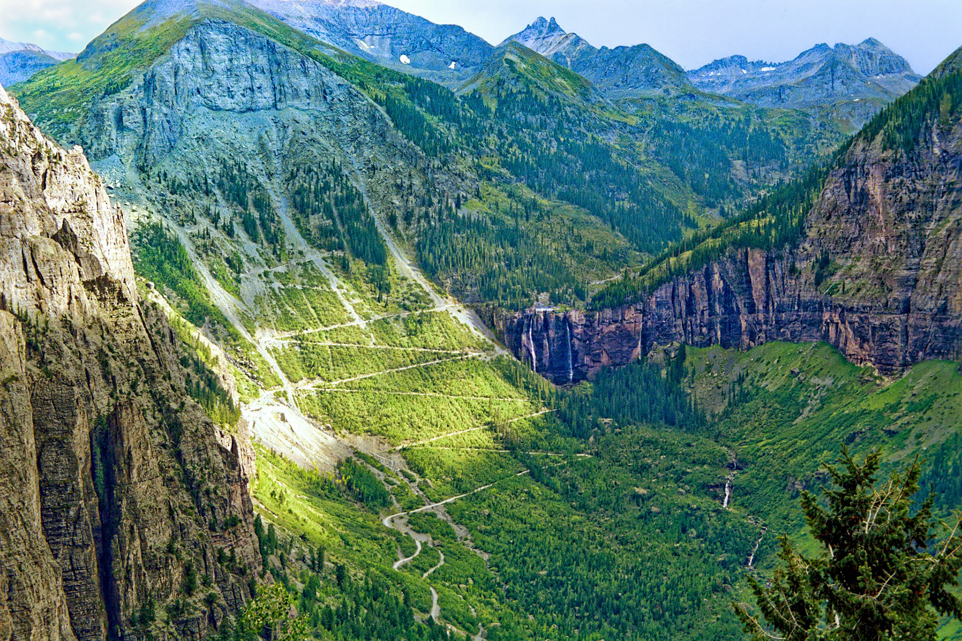 Sunlight pierces some clouds and highlights a road that switches back and forth while moving up the Ingram Peak (12,552 feet or 3,826 meters) massif. Near the center of the photo and just to the right of the switchbacks is Bridal Veil Falls, which are situated at 10,279 feet or 3,133 meters above sea level and at the head of a box canyon above Telluride, Colorado. Looking South-Southeast, this photo was taken from a vantage point near tree line on Tomboy Road, and it is about two miles (more than 3 kilometers) across the valley to the falls.