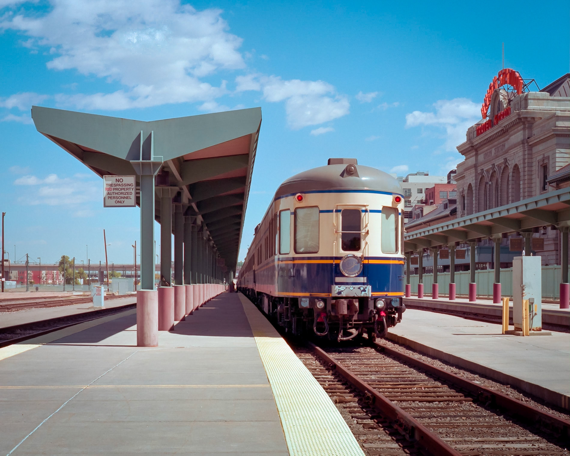 The California Zephyr, ready for boarding and Eastward-bound, sits on a track alongside the Union Station in Denver Colorado. With a familiar orange “Union Station, Travel by Train” sign on both the railyard and public entrance sides  advertising its primary purpose, the great hall, which was built in 1914, still functions as ticketing and check-in location for Amtrak. The site is now owned by RTD Denver, and it underwent a complete renovation in 2012 (about five years after this photo was made) as a transit oriented center for buses, light rail, and passenger trains. The remodeled Union Station complex also hosts a hotel, restaurants, and retailers serving both travelers and the general public.
