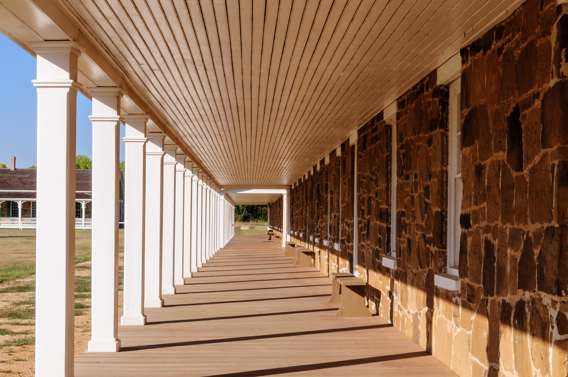 The rising sun casts shadows on the portico of the enlisted men's barracks located on the Northwest side of the parade grounds at the Fort Larned National Historic Site, which is  located along the Santa Fe Trail and near the City of Larned, Kansas. Officers' Quarters are seen in the background at center-left of the photograph.