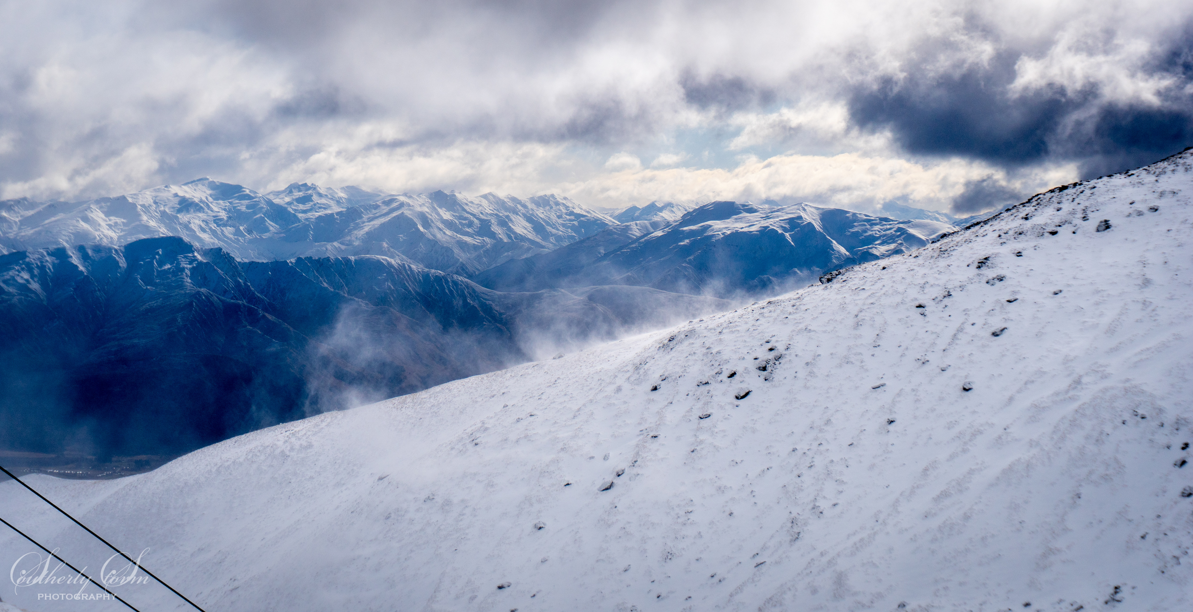 Wind blowing snow over the ridge, visible distant mountains beneath the cloud