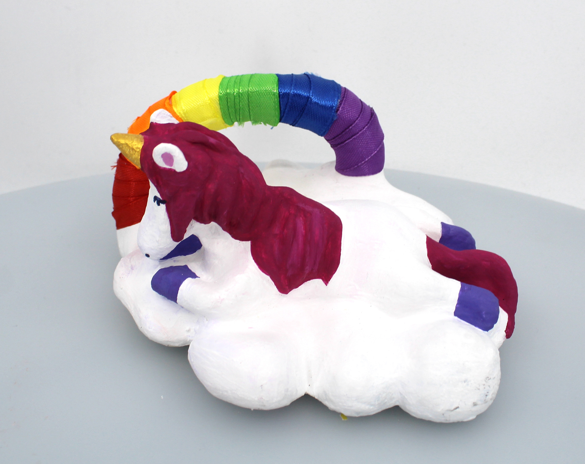 Oliver Cambroni This piece is an air dry clay sculpted unicorn laying on a cloud adorned with acrylic paint, featuring a rainbow crafted from various colors of ribbon. Its whimsical design captures the essence of fantasy and imagination, blending tactile craftsmanship with vibrant hues. The combination of clay, paint, and ribbon creates a charming and enchanting work of art sure to delight admirers of magical creatures and colorful creations.