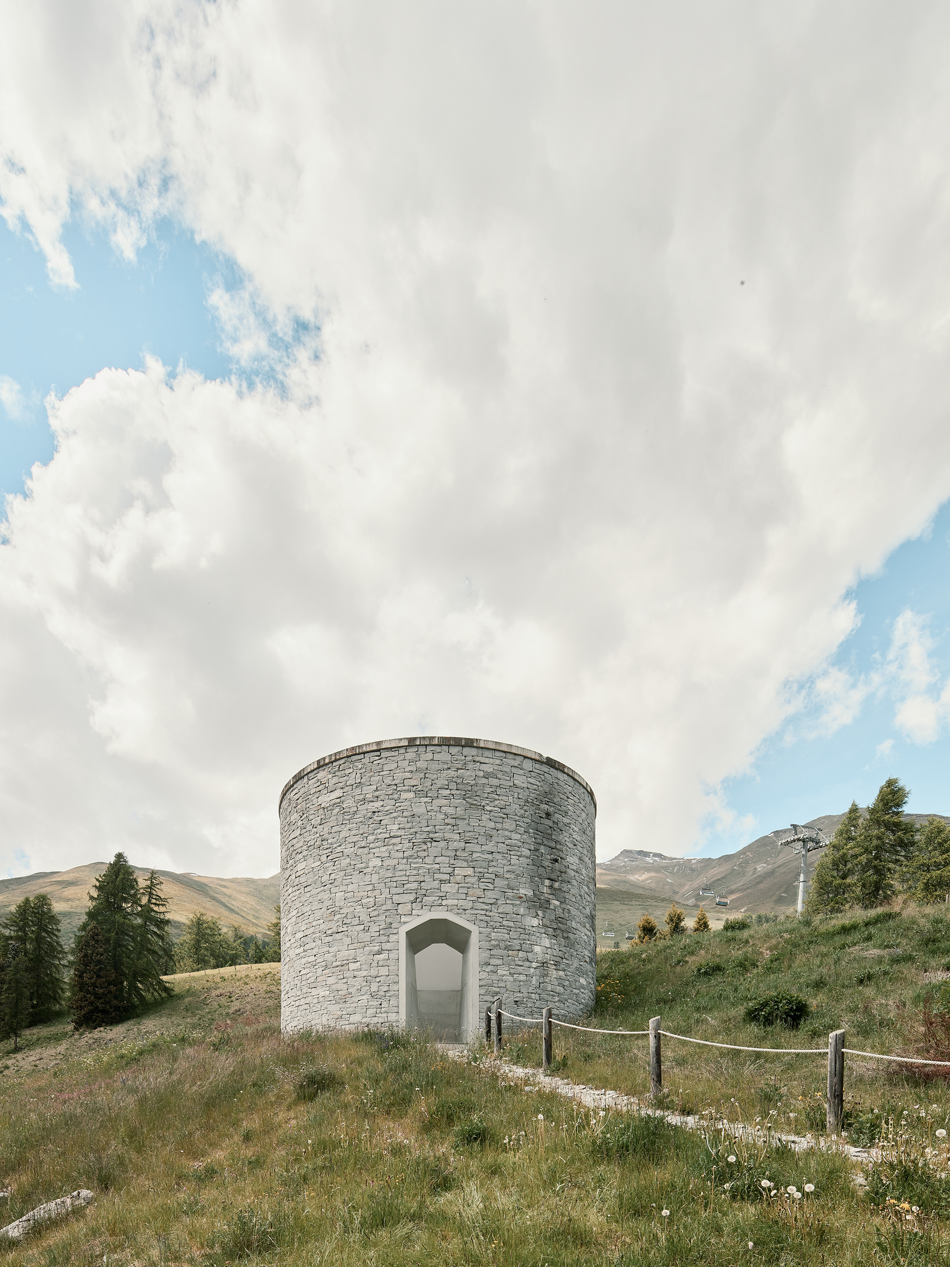 Outside view of James Turrell's Skyspace right next to Hotel Castell in Zuoz, Switzerland. Left and right of the structure are some trees, to the right is the ski slope which is out of service as the image is taken in June around mid day in sunny conditions and broad daylight. The landscape fills the lower third of the image whereas the upper two thirds are all sky.