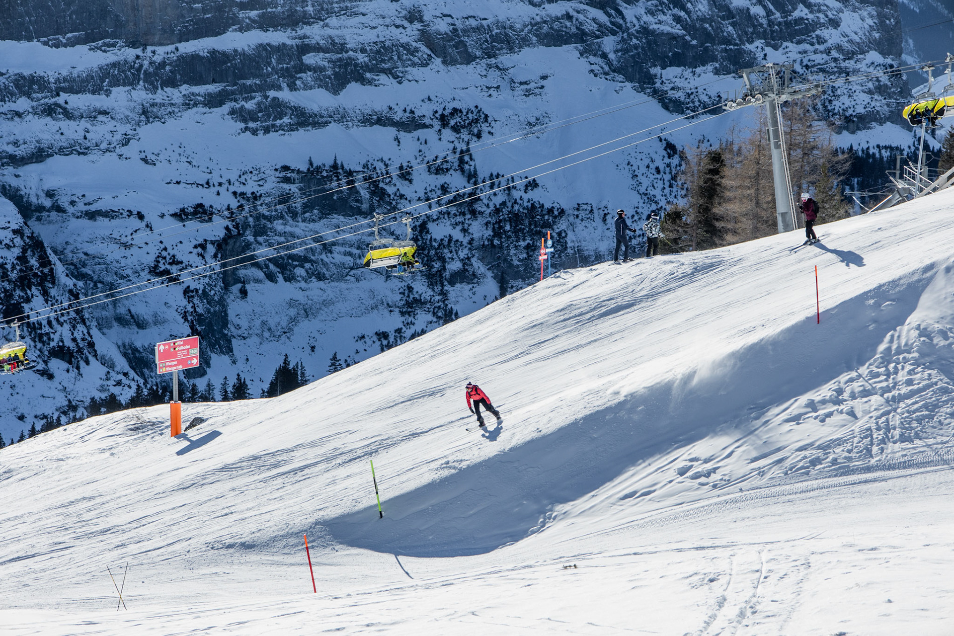 A man dressed in orange jacket is skiing down the hill. On the background are the cable cars and snowy mountains.