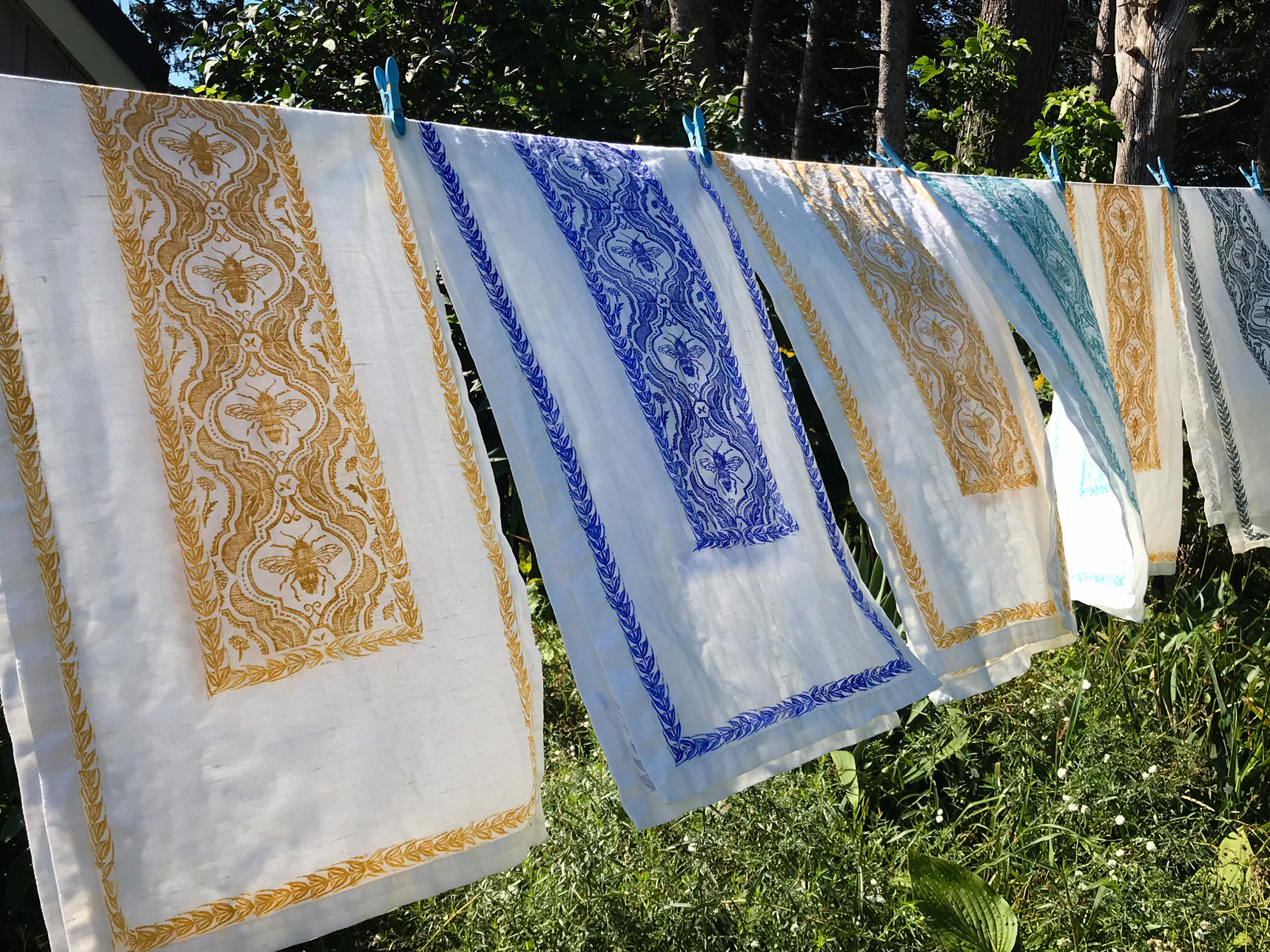 Bee table runners in various colours hanging on a clothes line. These items are hand printed by Canadian artist and printmaker Kristine MacGregor