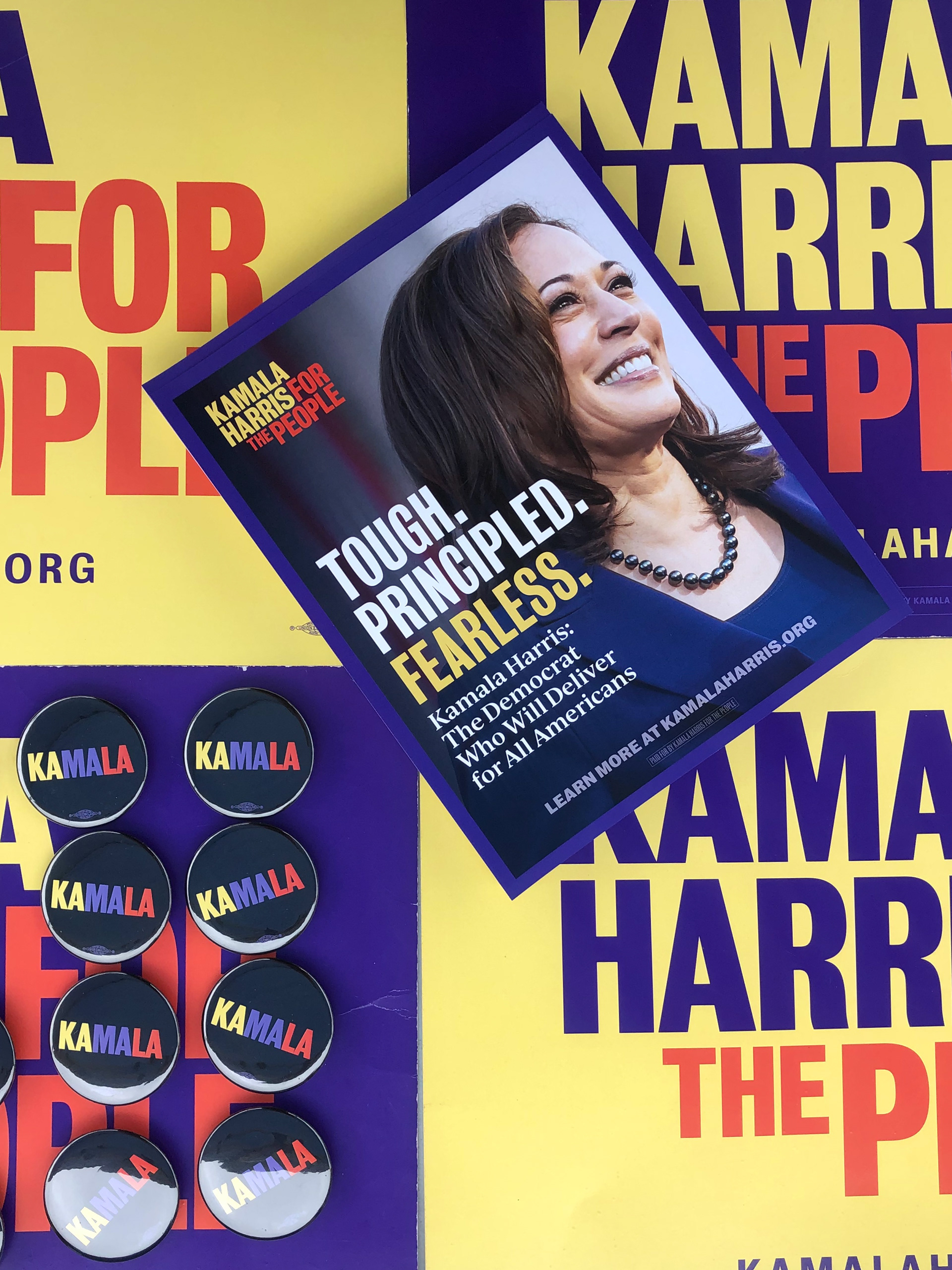 Kamala Harris Flyer reading "TOUGH. PRINCIPLED. FEARLESS." lying diagonally across alternating purple and yellow "Kamala Harris for the People" sign with small black "KAMALA" buttons in the corner