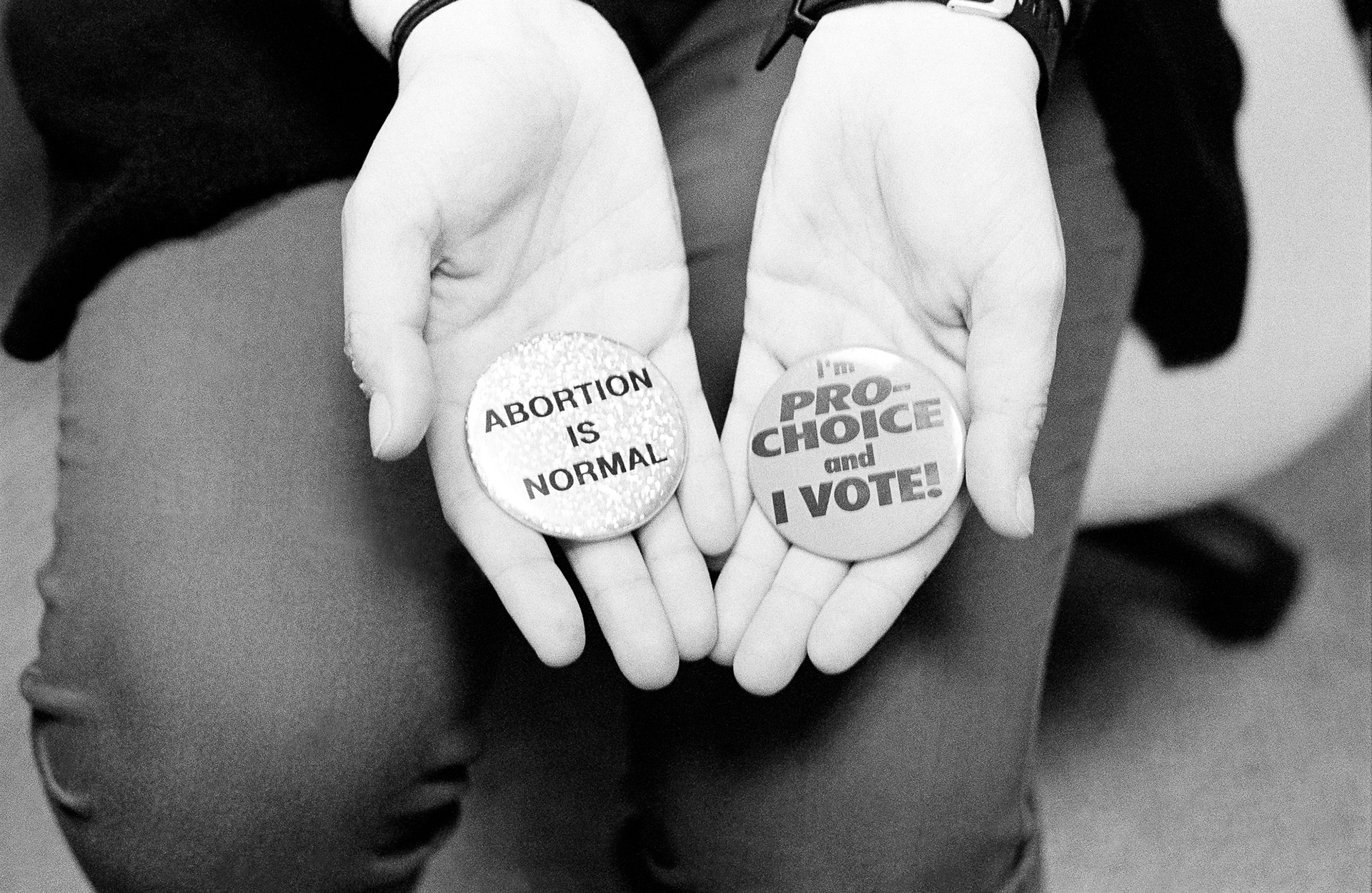 A woman's hands holding two buttons - one that reads "ABORTION IS NORMAL", the other "I'm PRO-CHOICE and I VOTE!"