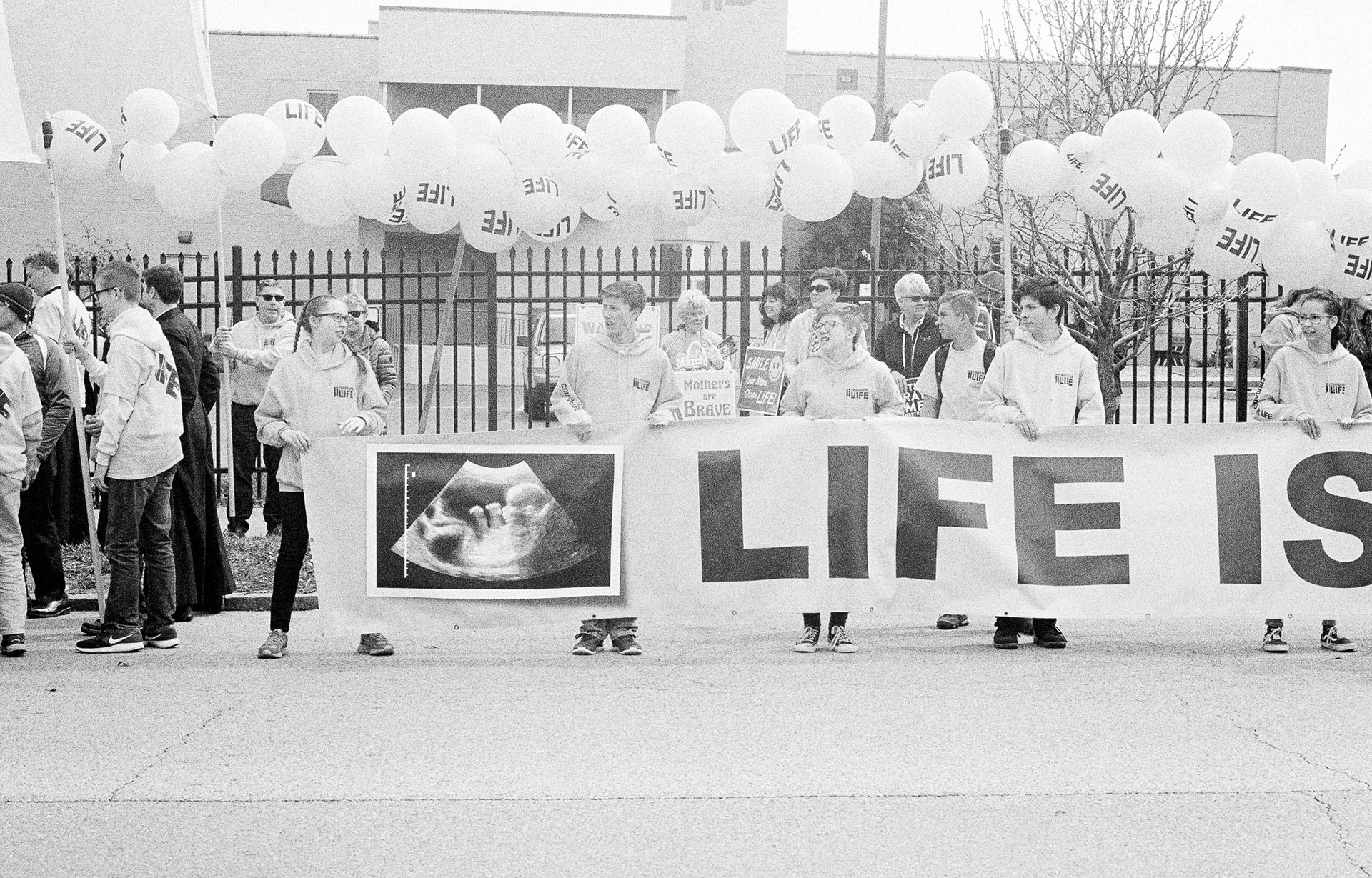 teenagers in front of a fence adorned with balloons that say "LIFE", holding a cut-off banner with an image of a sonogram and text that reads "LIFE IS..."