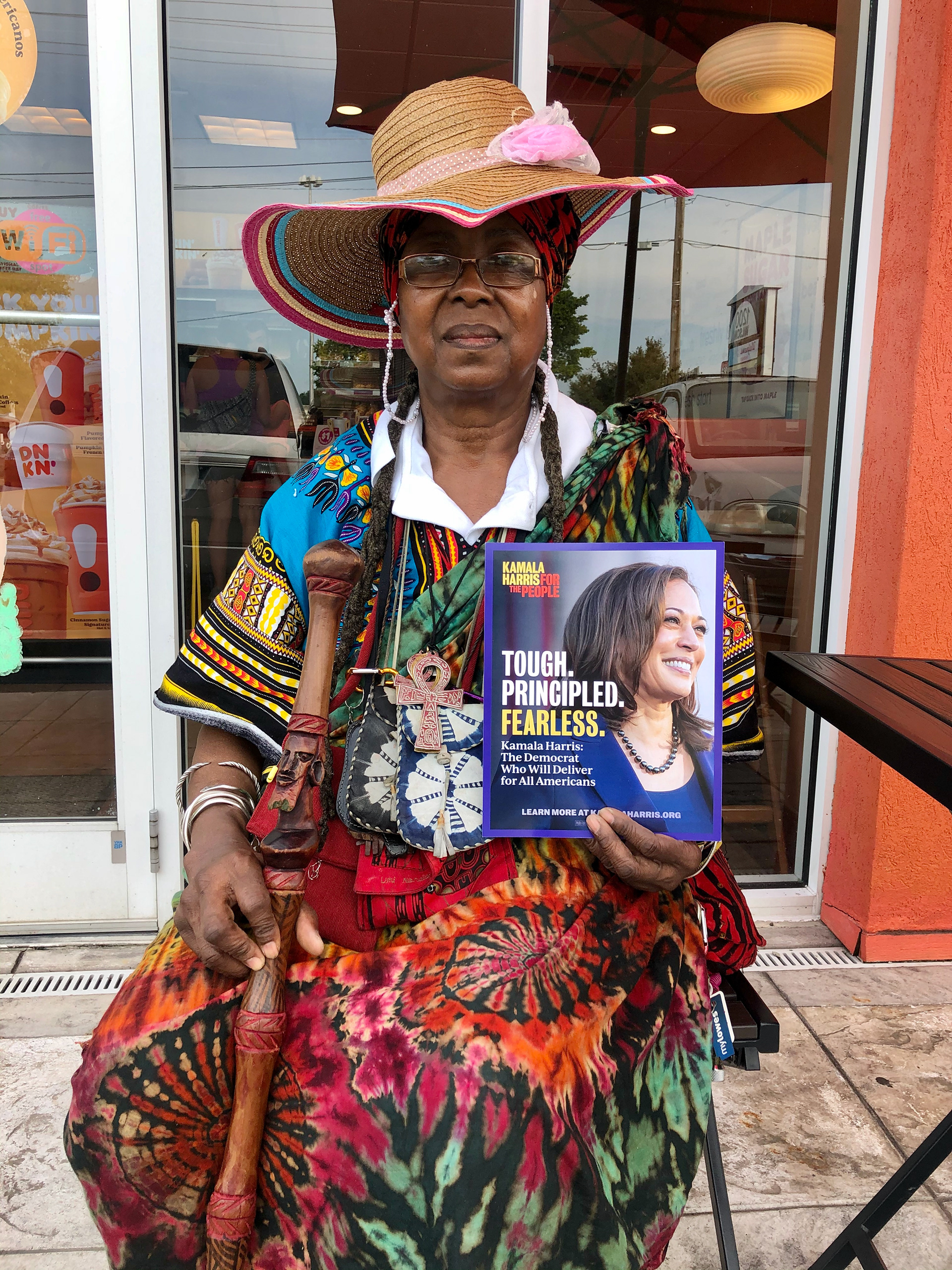 Photograph of an older black woman, seated, in colorful tribal dress, a large hate, and a cane. She is holding up a Kamala Harris flyer that reads "Tough. Principled. Fearless."
