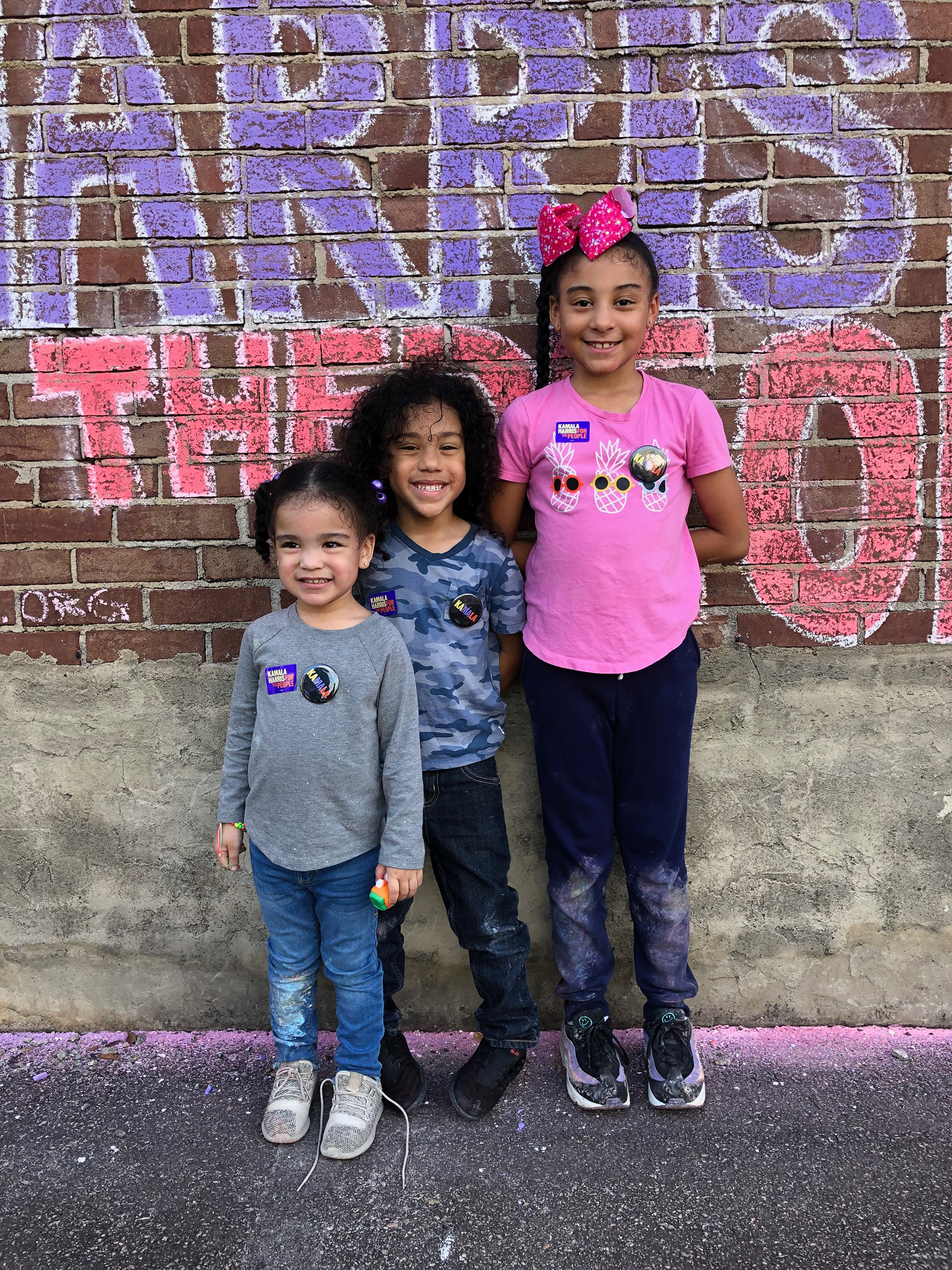 Three children, ordered left to right from shortest to tallest, stand smiling in front of a red brick wall that reads "HARRIS THE PEOPLE" with the rest of the writint cut