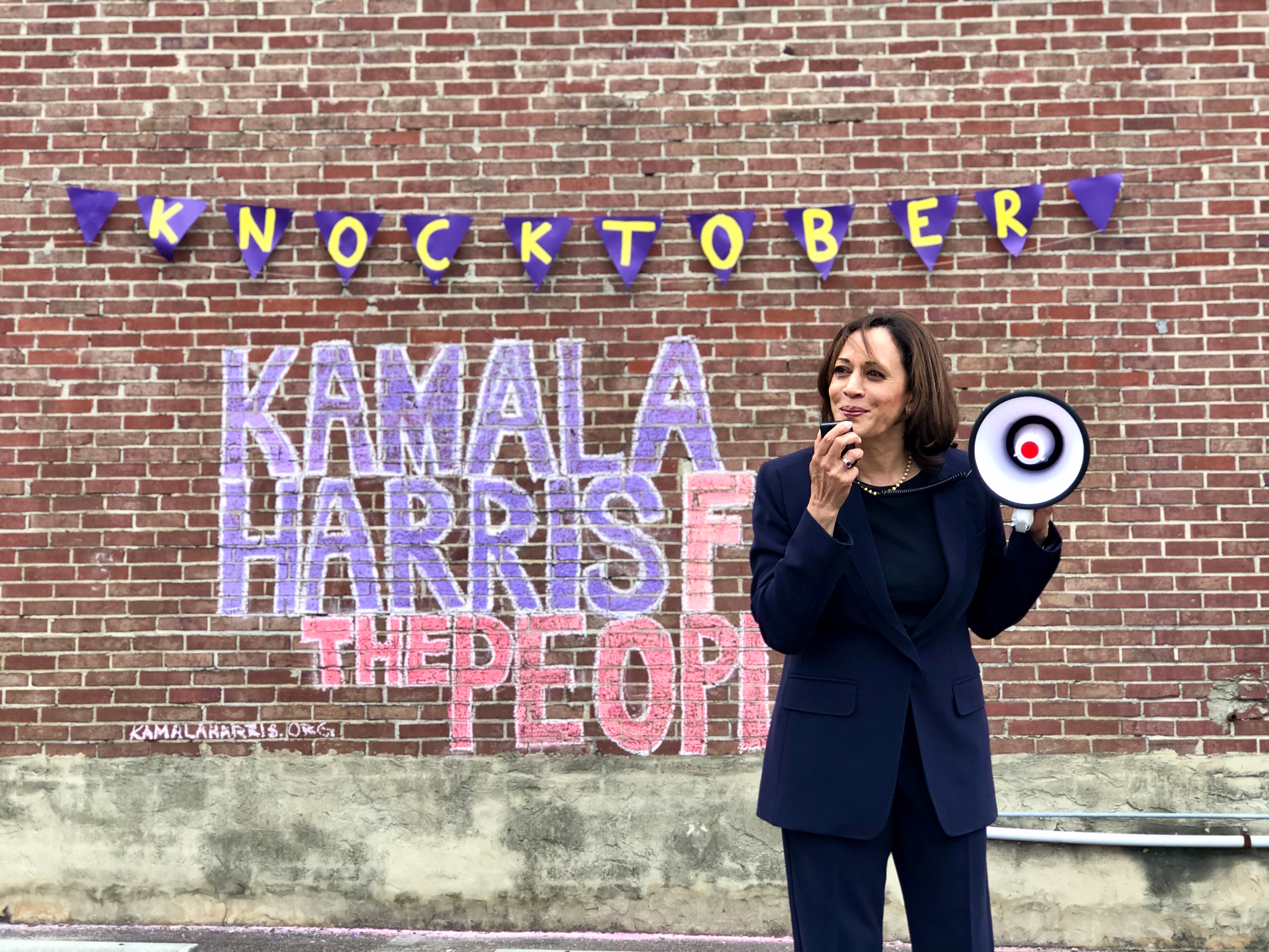 Kamala Harris holding a small megaphone, smiling off-camera, in front of a red brick wall with a sign that says "KNOCKTOBER" and "Kamala Harris For the People" drawn in purple and red chalk