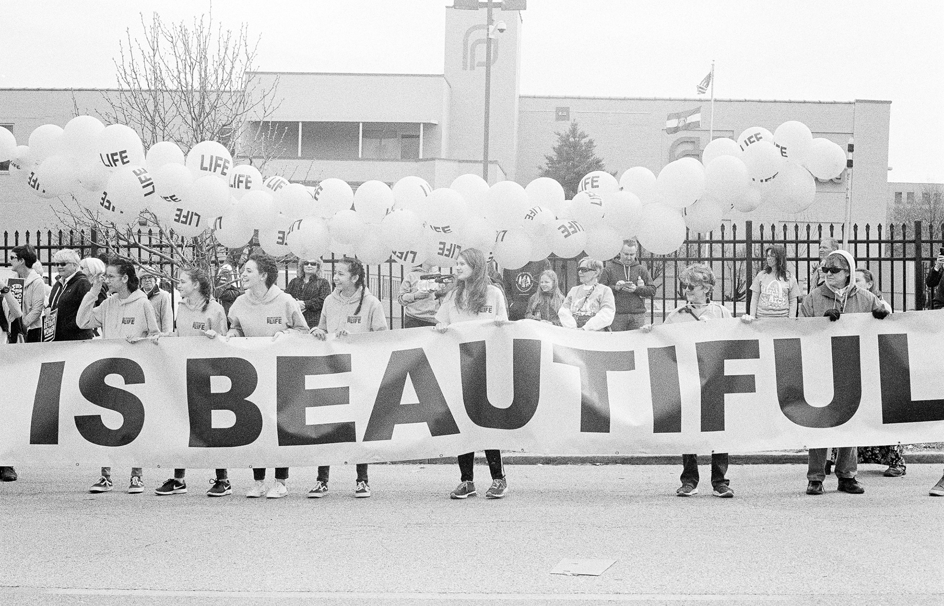 teenagers in front of a fence adorned with balloons that say "LIFE", holding a cut-off banner that reads "...IS BEAUTIFUL"