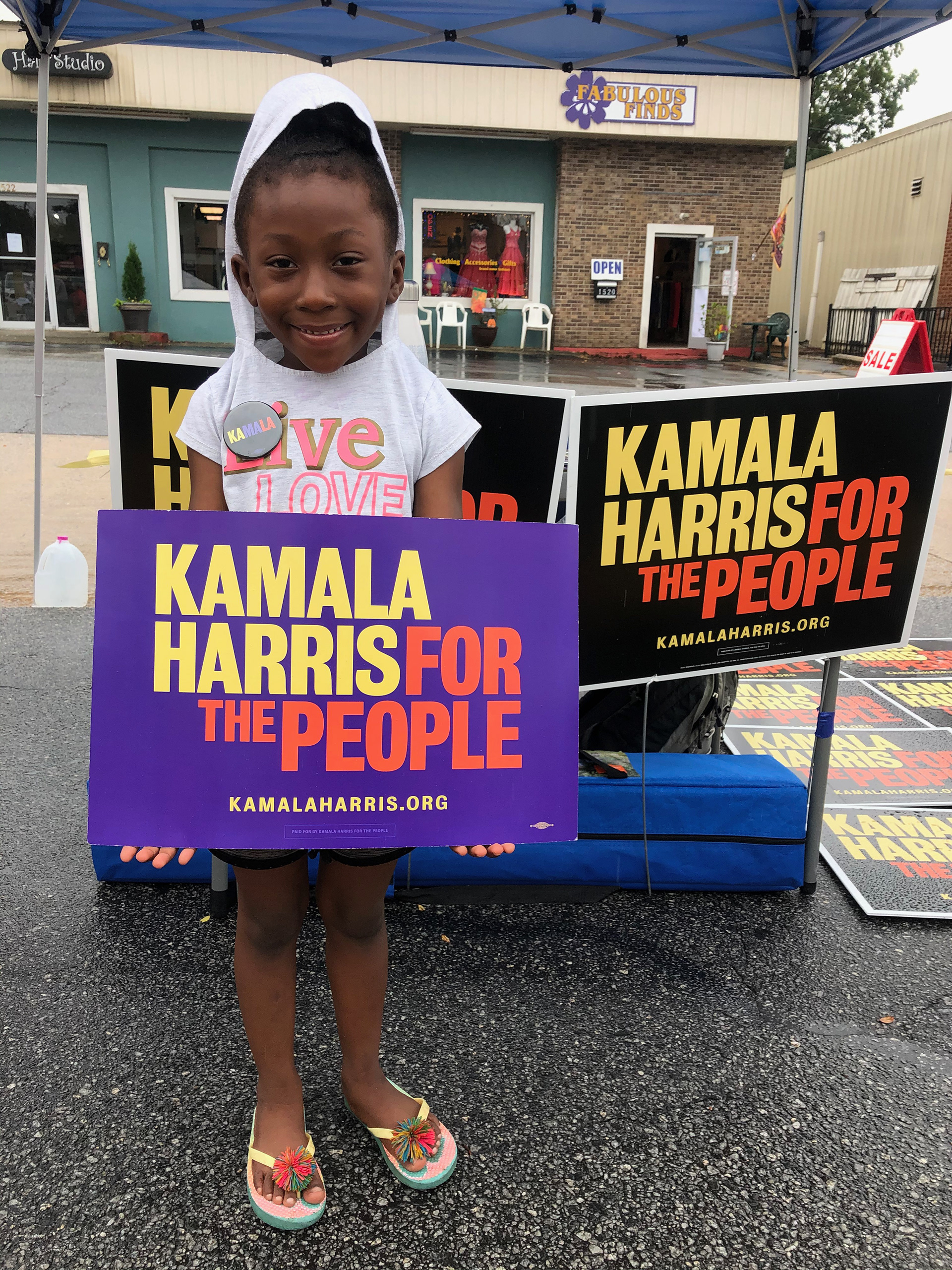 Small black child wearing a gray t-shirt with hood over her hair and a "KAMALA" pin, smiling and holding up a purple "Kamala Harris For the People" sign,. In the background, there are several black yard signs saying the same thing and several stores.