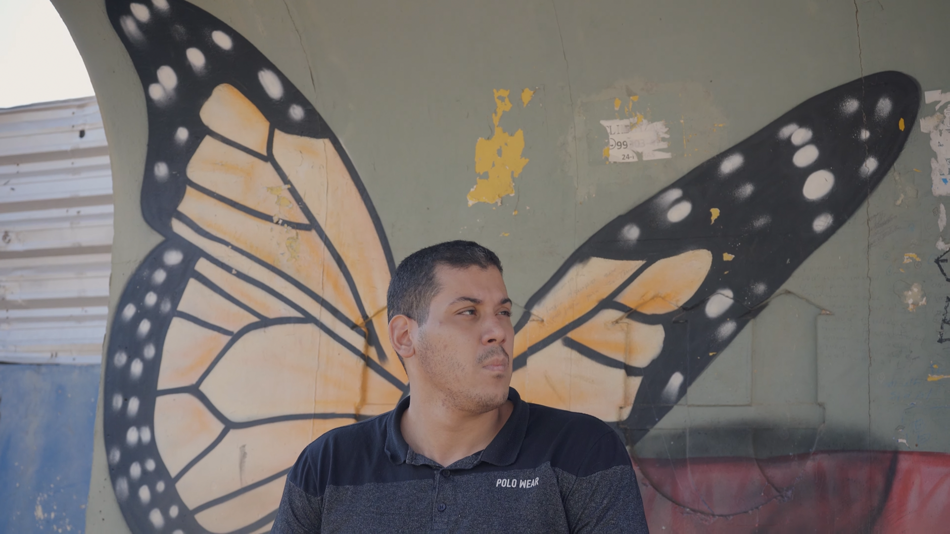 Foto do filme, um homem jovem de cabelo curto, de camiseta polo cinza, olha concentrado para a direita; no fundo, uma parede de concreto com um grafite grande de asa de borboleta, amarela e preta, que parece sair das costas do homem