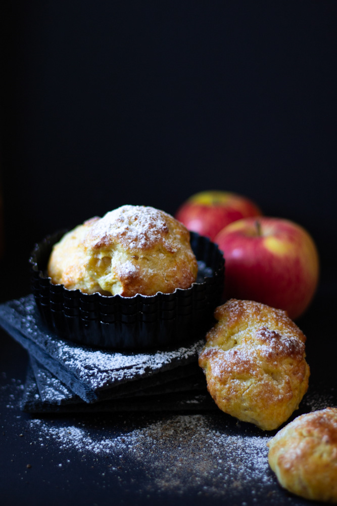 Mini apple pies on black ramekins on black background and red apples further away. A lot of icing sugar and black square plates, piled onto each other