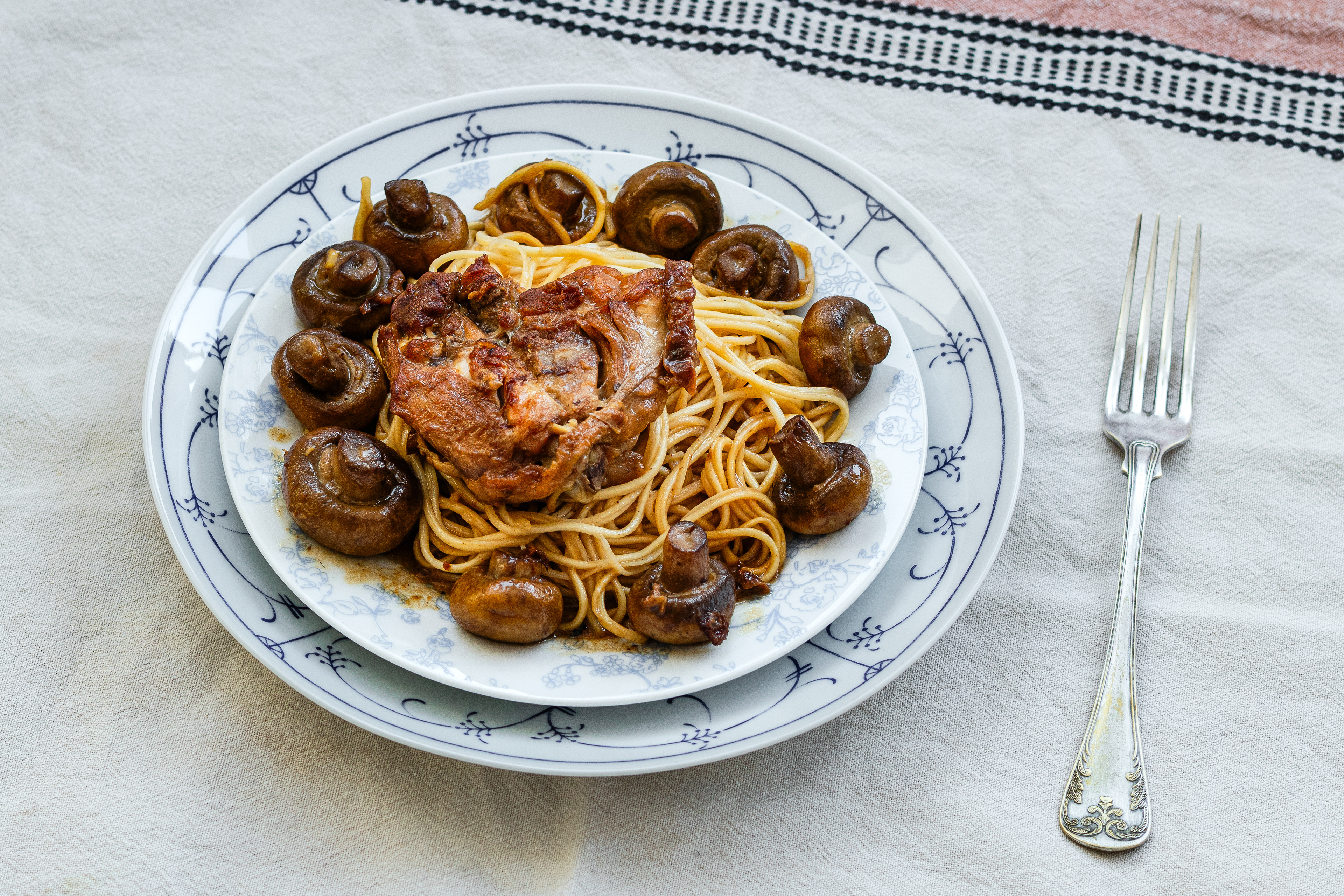 Rustic food photograph of spaghetti topped with braised meat and whole mushrooms, styled on a linen tablecloth