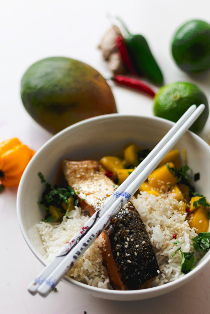 Grilled salmon with mango salsa, rice, and sesame seeds in a white bowl, with ceramical chopsticks and colorful fruit and veggies aside.