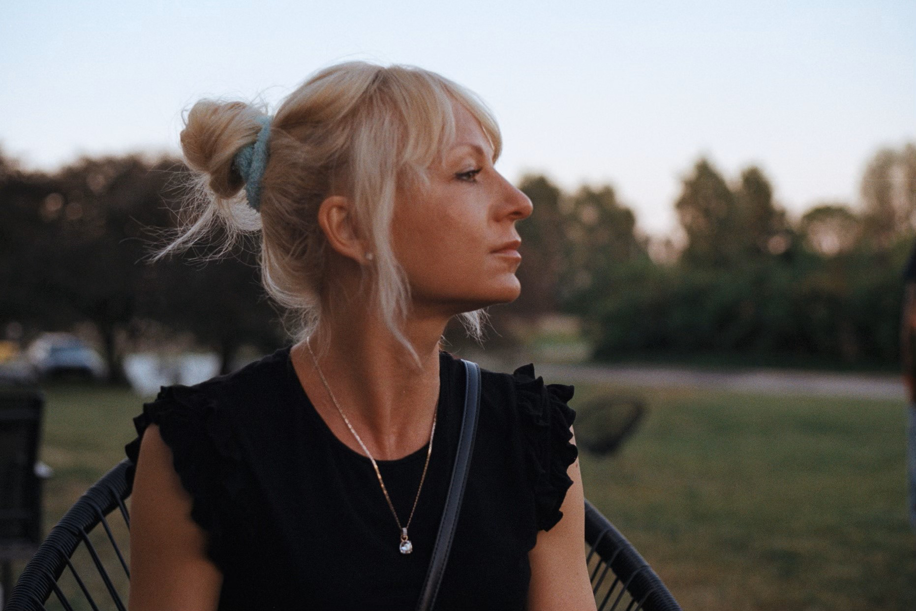 Slightly vintage style edited photo in smooth sunset colors,. A sun-tanned girl looking to the left, so that only her profile is visible. Her hair are a bit curly, blond and tied into a bun. The light is smooth and leaves an impression of a summer night. There is a lot of dark green field and trees as a background. The girl is wearing black top and she has a golden necklace and a string of a handbag is visible over all. She's sitting on a chair, but only the upper part of her body is captured (a head shot)