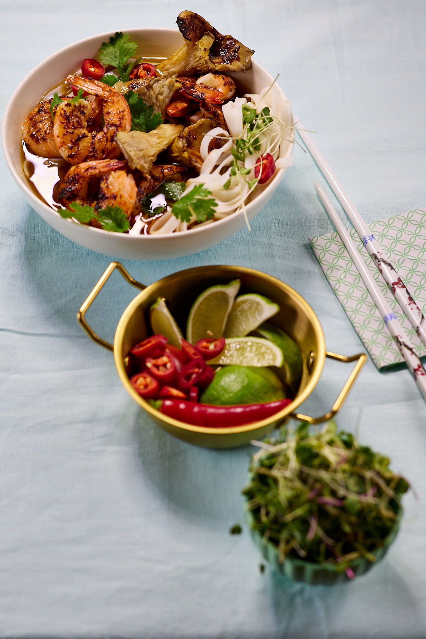 Spicy prawn soup with rice noodles, Oyster mushrooms, and lime wedges, garnished with fresh herbs and chili peppers and micro greens - garnishes placed into 2 additional separate bowls. Ceramic white chopsticks with some decorative pattenrs and a white paper with light gren patterns underneath them. Everythin is place diagonally on a light blue table cloth