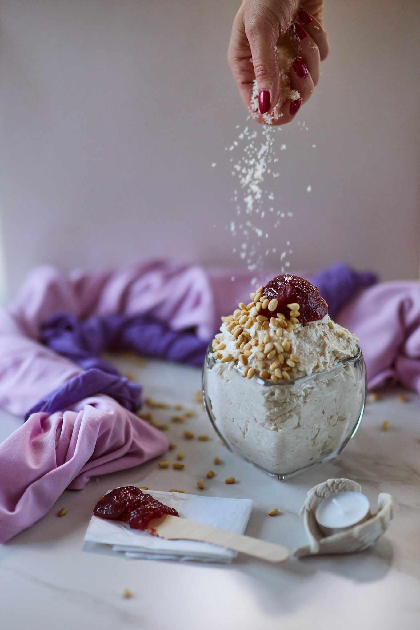 Woman's hand with red nails sprinkling shredded coconut over a white mpusse dessert dish in glass bowl, topped with pine nuts and red jam. 2 tones of purple towels on the background, a small candle and a wooden knife with the jam laying on white marble surface and a piece of white paper.
