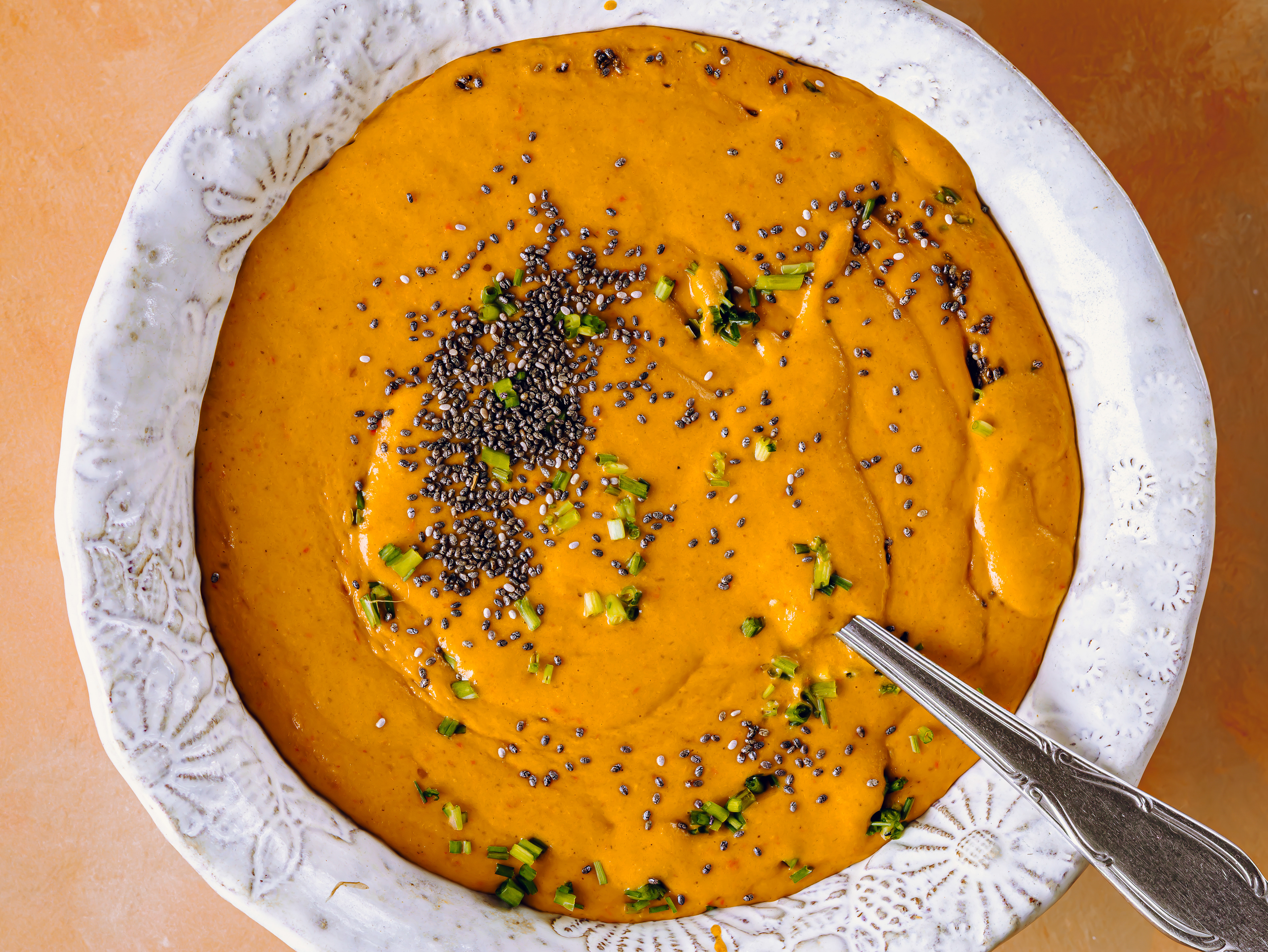 High-angle flat lay food photography of a rich orange puree soup garnished with fresh herbs and seeds. Shot in bright, soft light that highlights the rich textures of the food, the detailed ceramic bowl, and the warm-toned backdrop.