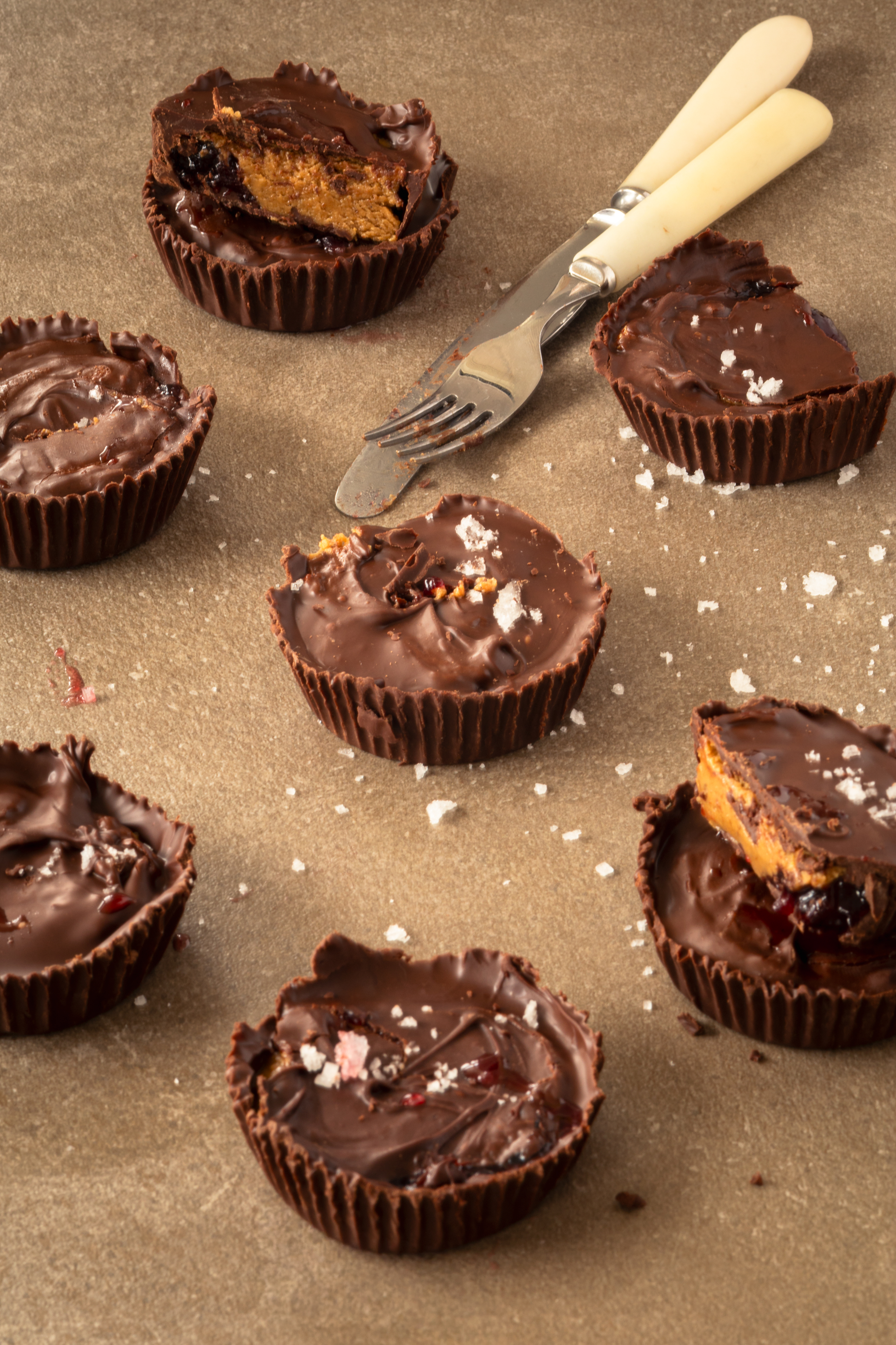 Homemade dark chocolate sunflower butter cups scattered on a surface, some cut open to reveal seed butter and jam filling, sprinkled with flaky sea salt and served with a small fork