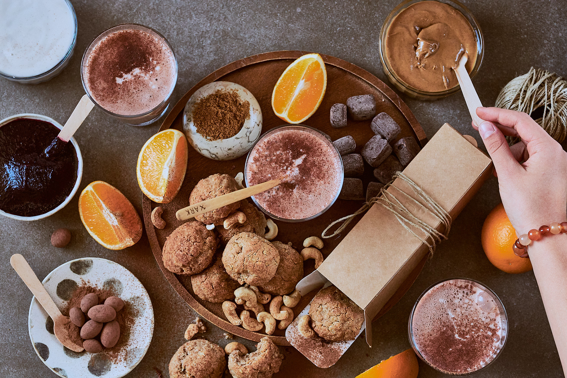 Variety of warm chocolate treats, including hot chocolate, cookies, and chocolate candies and truffles, almonds and cashews, jam and coconut condensed milk, presented with oranges and other ingredients. Busy scene on a gray stone surface, a child's hand taking a spoonful from a brown nut butter. Cookies are partially falling out from a box, which is tied with a rustic tie..