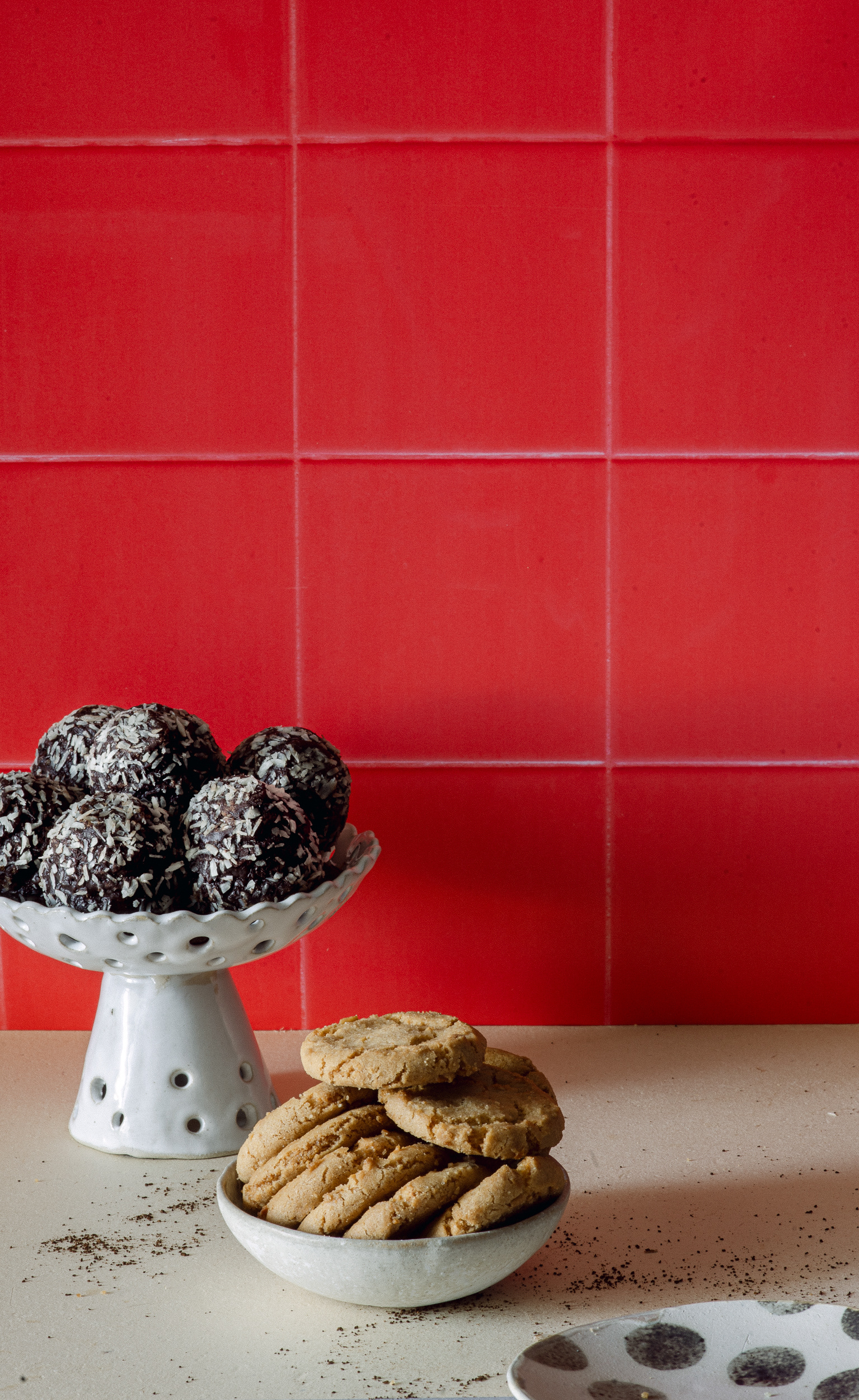 Chocolate coconut energy balls on small white tall serving plate, cookies in a bowl on an old book. Coffee crumbs, a small plate with dots, everything placed diagonally next to a bright red wall