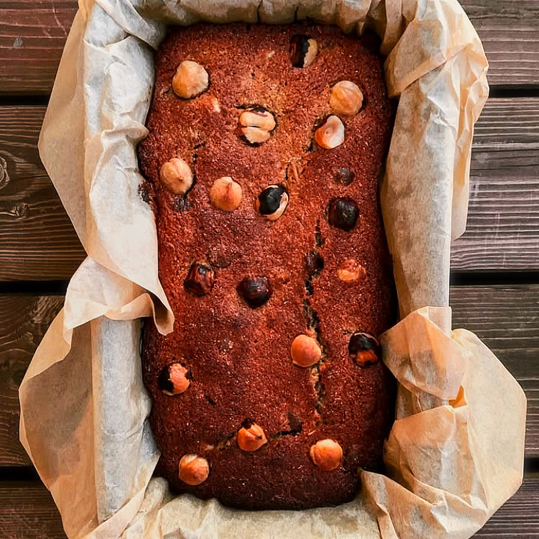 A bread loaf in a tin, baking paper edges reaching out of the tin and the bread is covered with roasted hazelnuts, the tin is on a rustic wooden floor