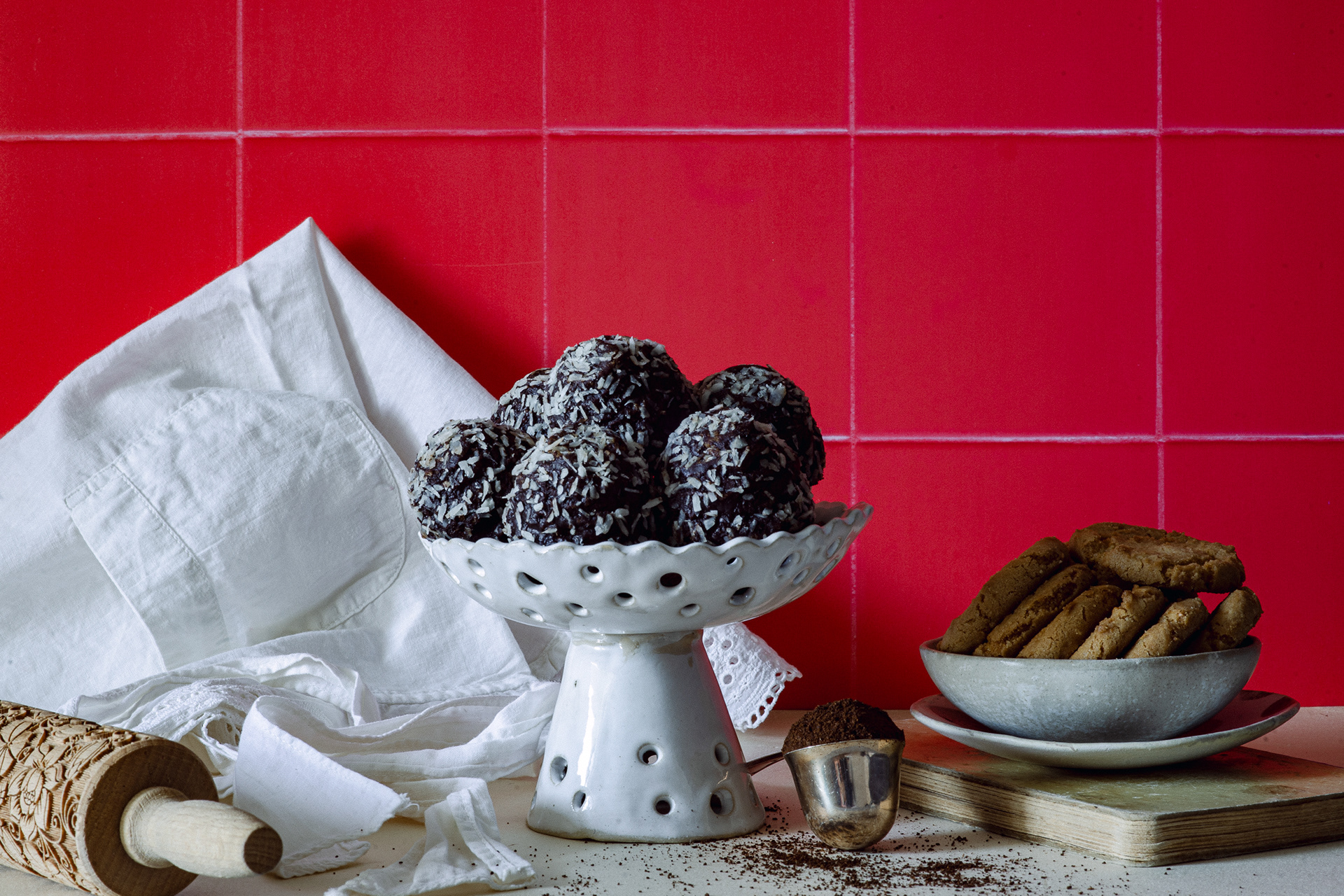 Chocolate coconut energy balls on small white tall serving plate, cookies in a bowl on an old book. Coffee crumbs, rolling pin and white apron next to a bright red wall