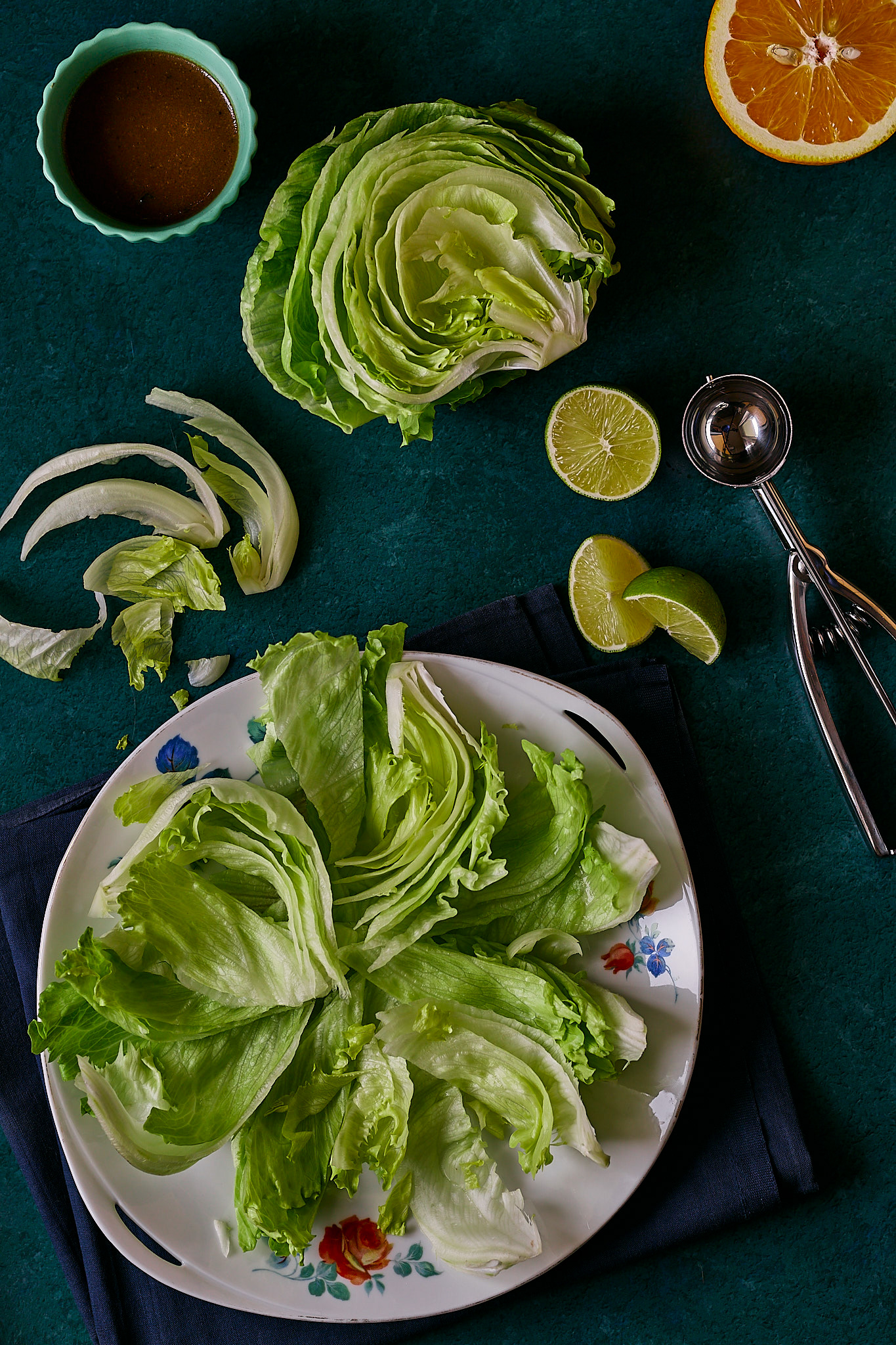 Freshly chopped lettuce leaves arranged on a patterned plate, alongside lime wedges, an orange slice, and a small bowl of dressing. Dark green backdrop.