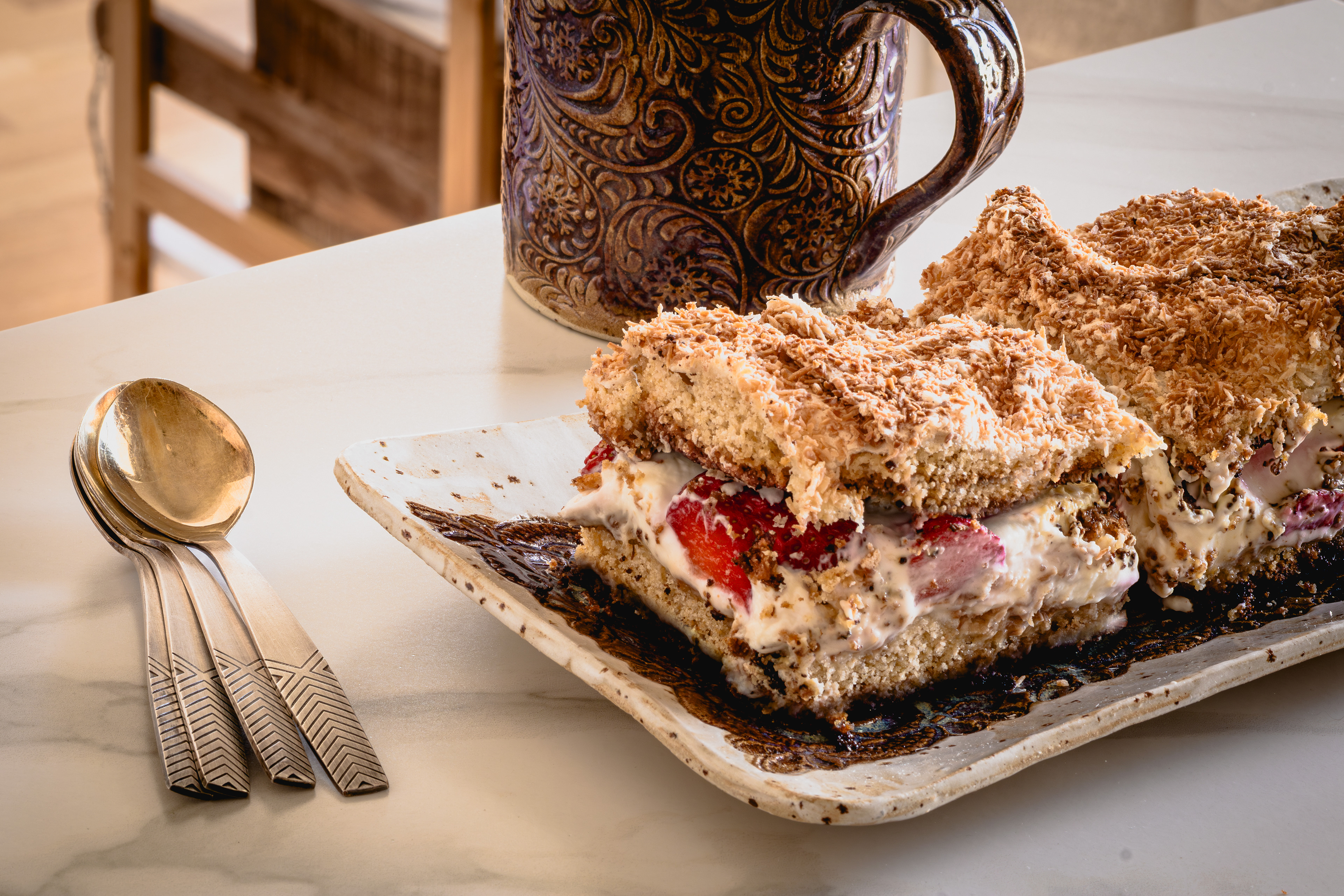 Layered strawberry cream meringue cake squares topped with toasted coconut on a ceramic serving plate beside vintage spoons and a patterned ceramic mug.