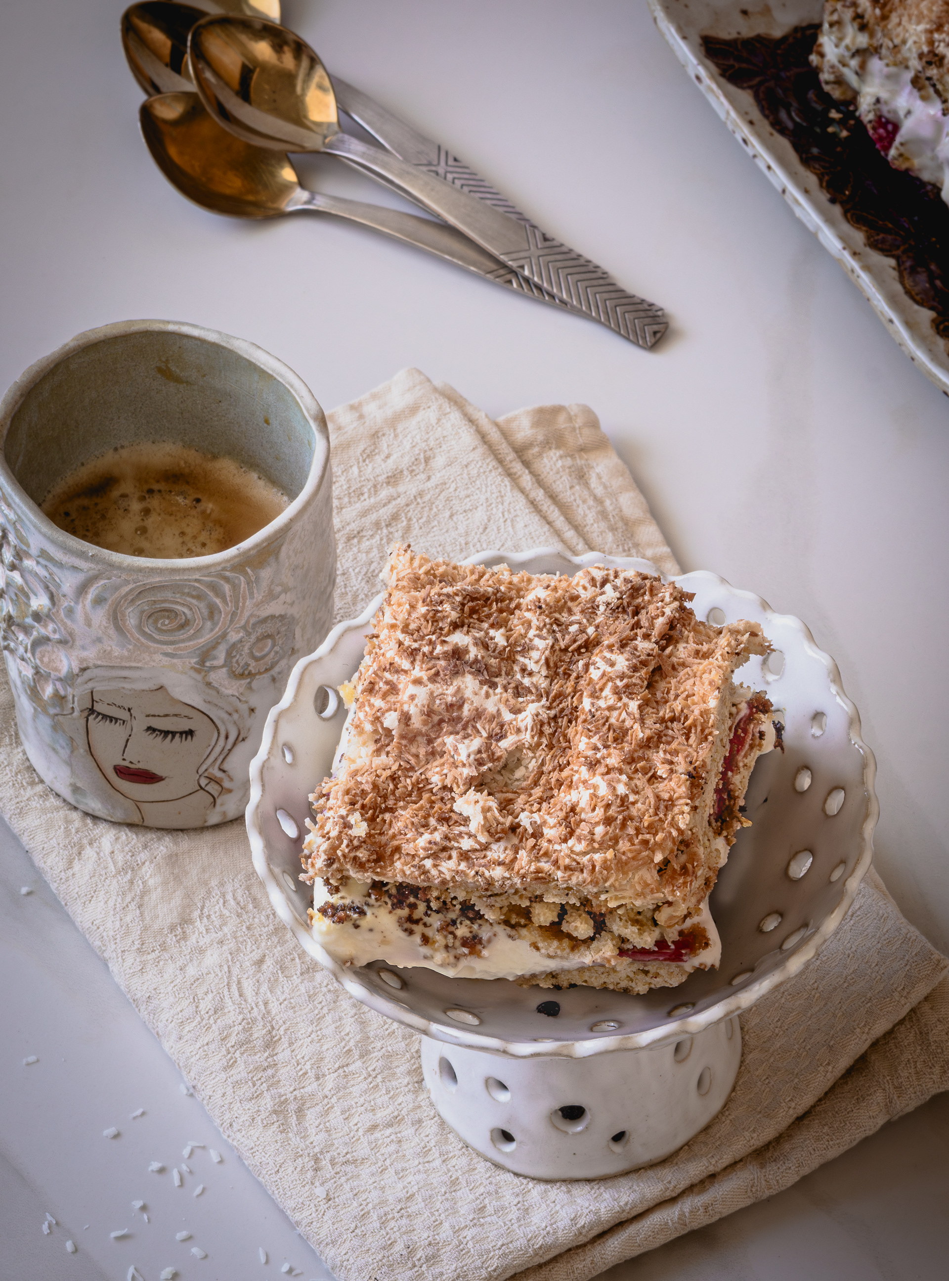 Layered strawberry cream meringue cake squares topped with toasted coconut on a ceramic serving plate beside vintage spoons and a patterned ceramic mug.