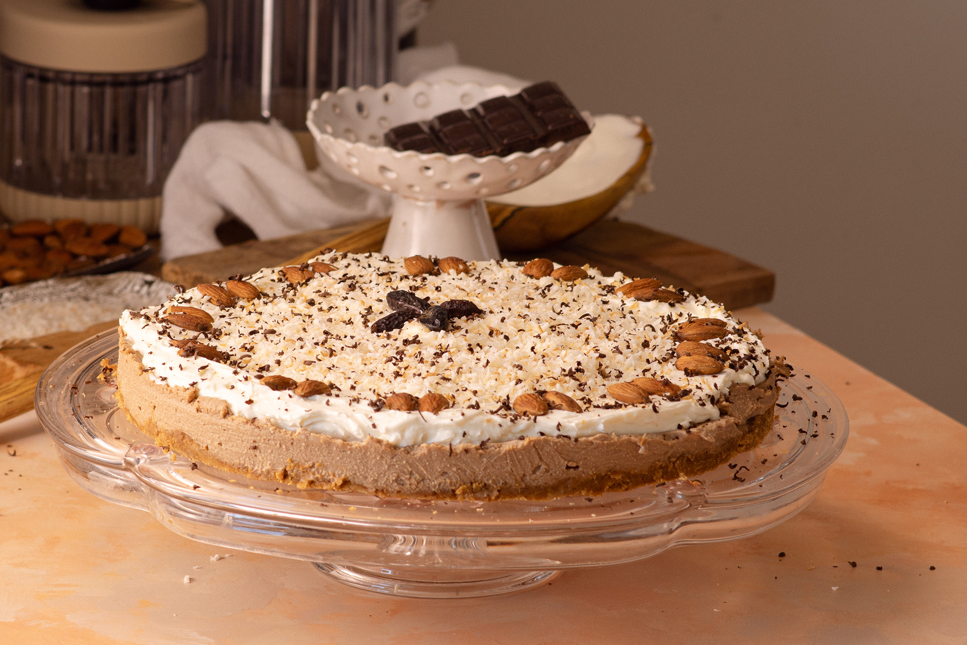 Chocolate Coffee No-Bake Cheesecake on a glass cakestand on orange and teal colored countertop. The cake is decorated with roasted almonds, slivered dark chocolate slivered and whole tonka beans and roasted shredded coconut 