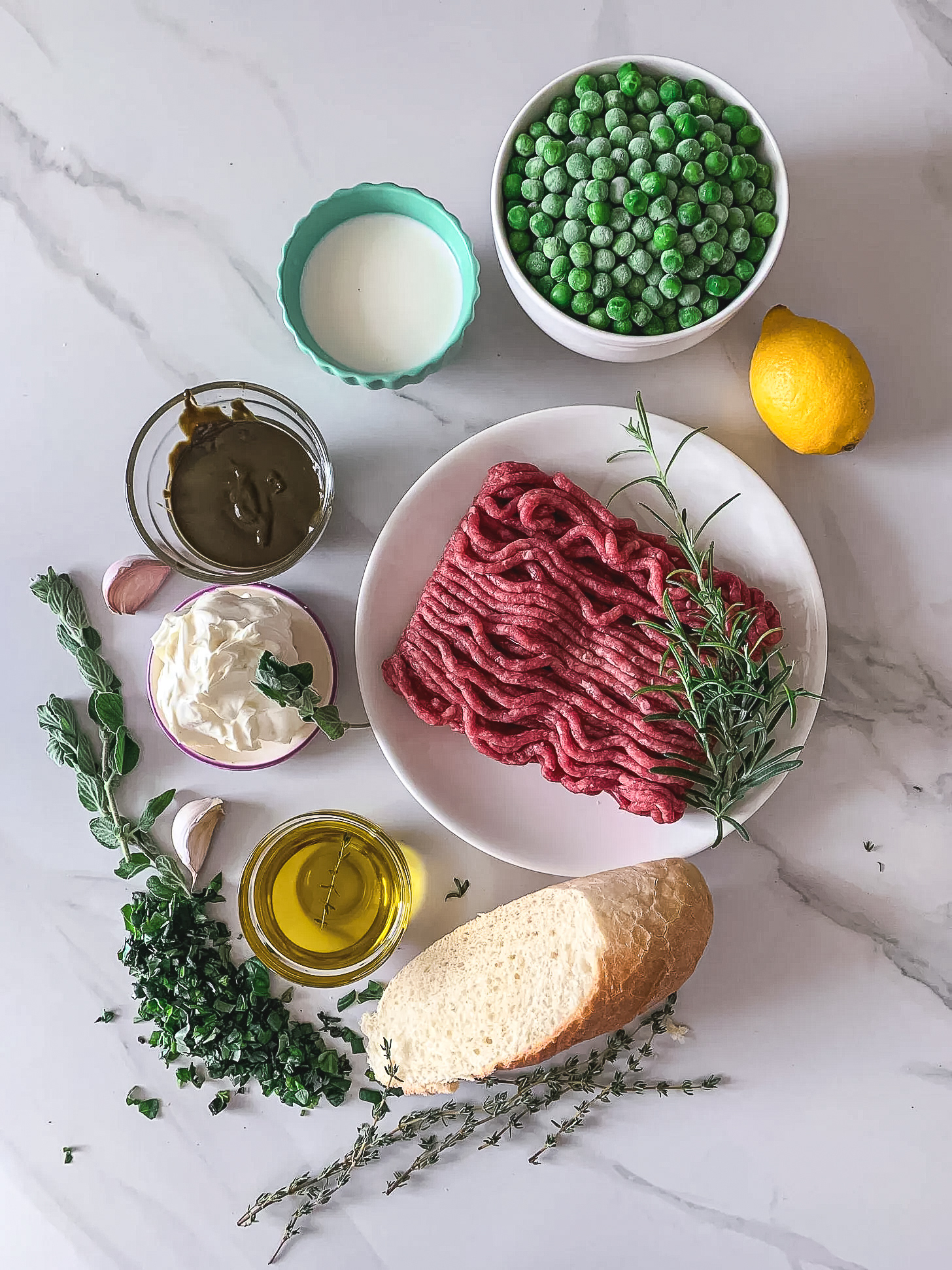 Ingredients for herby Greek meatballs with pea pistachio pesto, including ground beef, frozen peas, fresh herbs, garlic, olive oil, lemon, cream, and bread on a light marble surface.