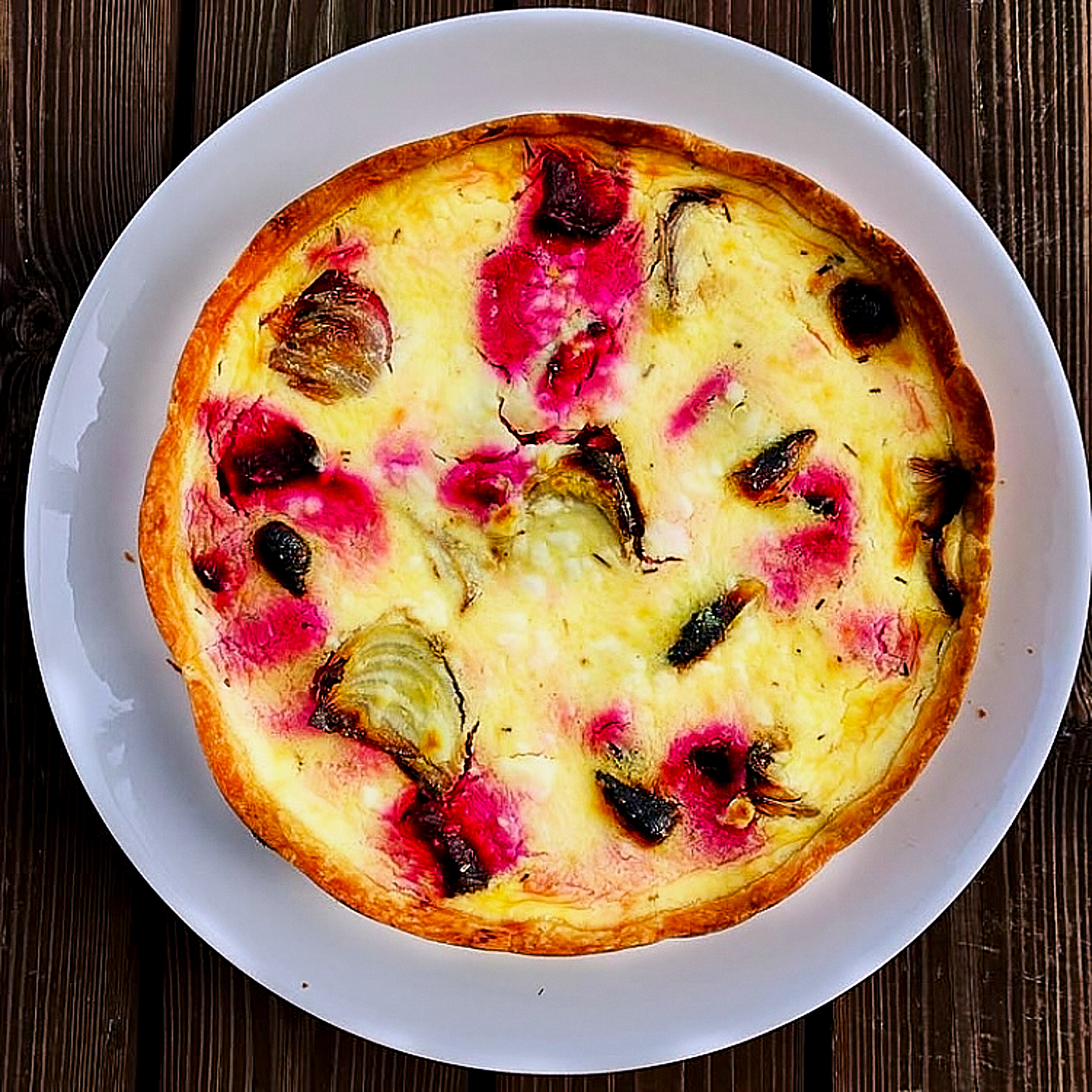 A colorful quiche with beetroots, char-grilled onions and Ricotta on a white plate and rustic wooden surface