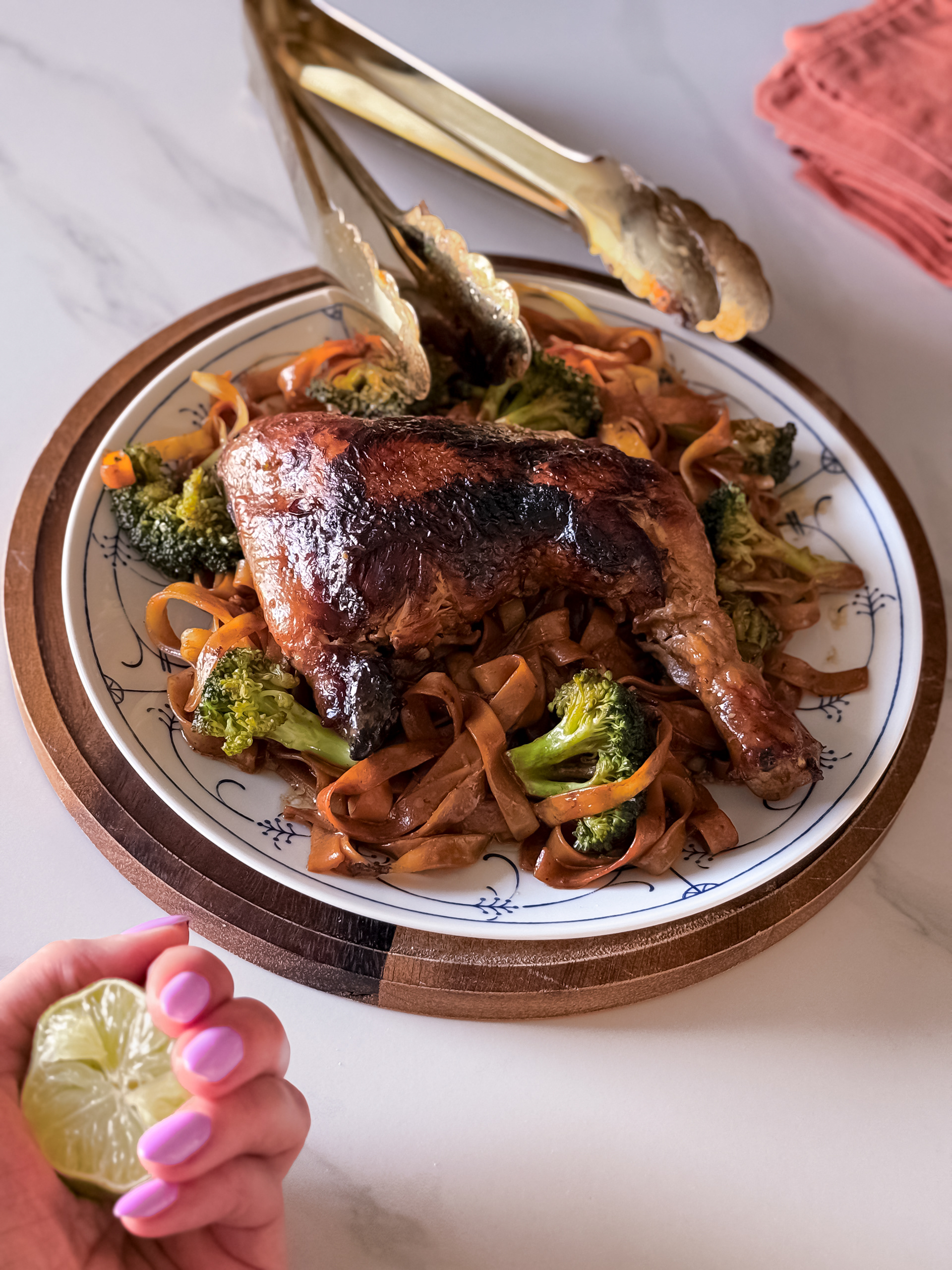 Dark grilled skin chicken leg on a pillow of brown teriyaki tagliatelle and broccoli. A lime halve and large golden tongs.