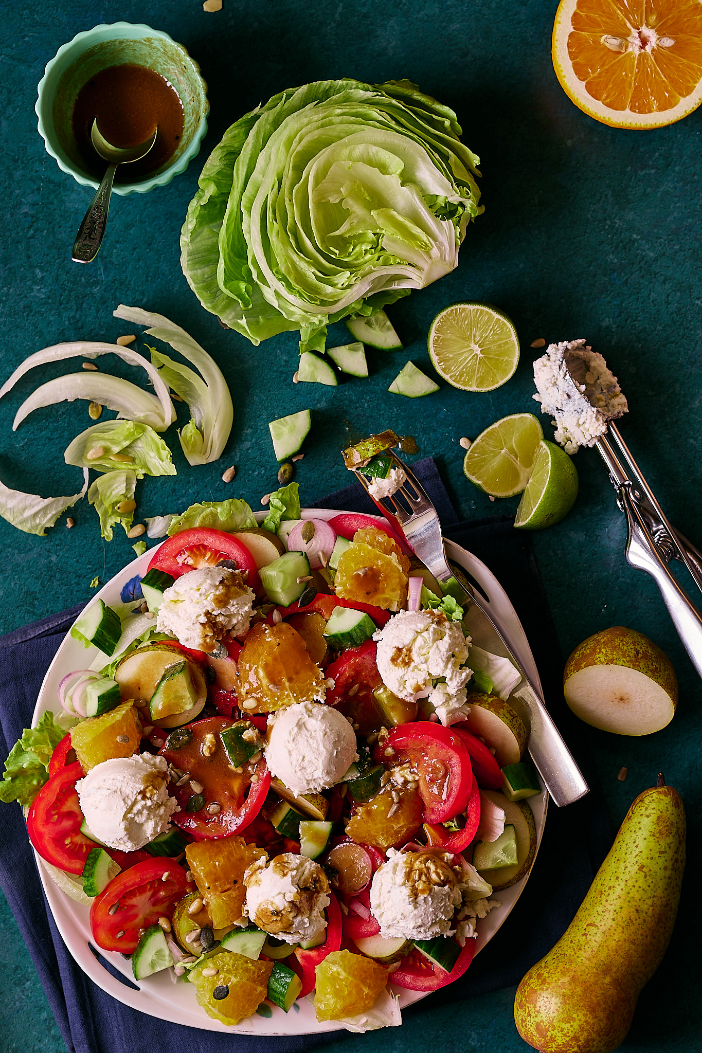 Fresh salad with mixed greens, tomatoes, cucumbers, oranges, pears, and feta cheese balls, drizzled with a dark vinaigrette, arranged on a white plate., placed on a dark blue-green backdrop