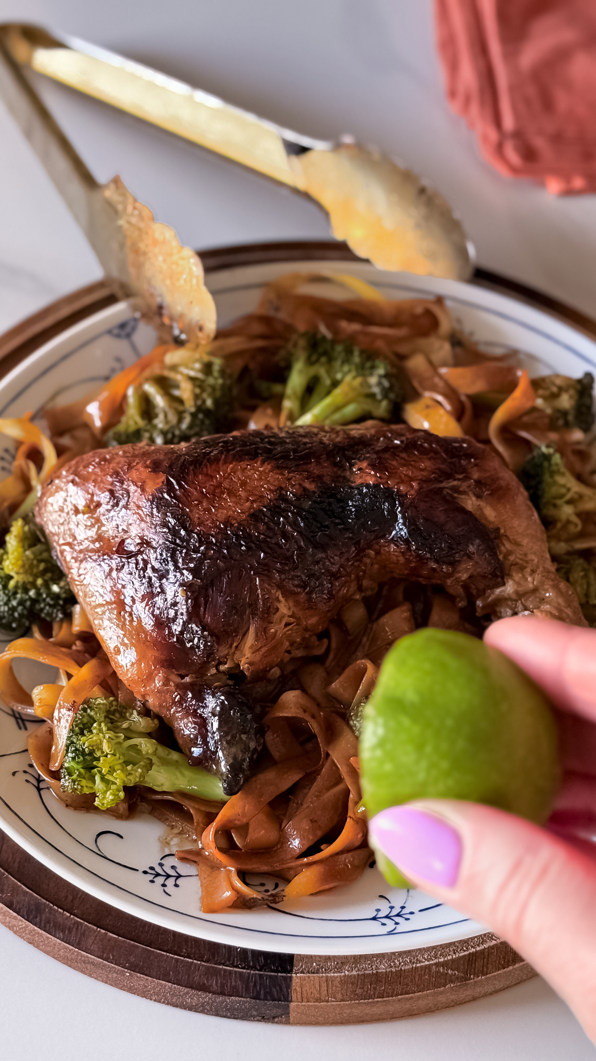 Dark grilled skin chicken leg on a pillow of brown teriyaki tagliatelle and broccoli. A lime halve and large golden tongs.