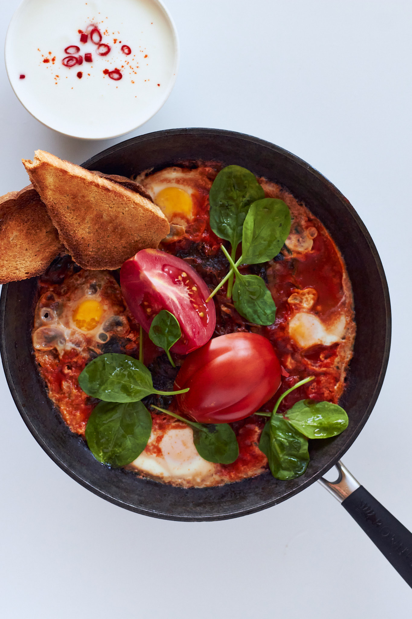 Savory breakfast dish: spinach shakshuka, made of fried eggs, tomatoes, spinach, and toast in a dark skillet. A bowl of Greek yogurt with chili slices on the side. White background.