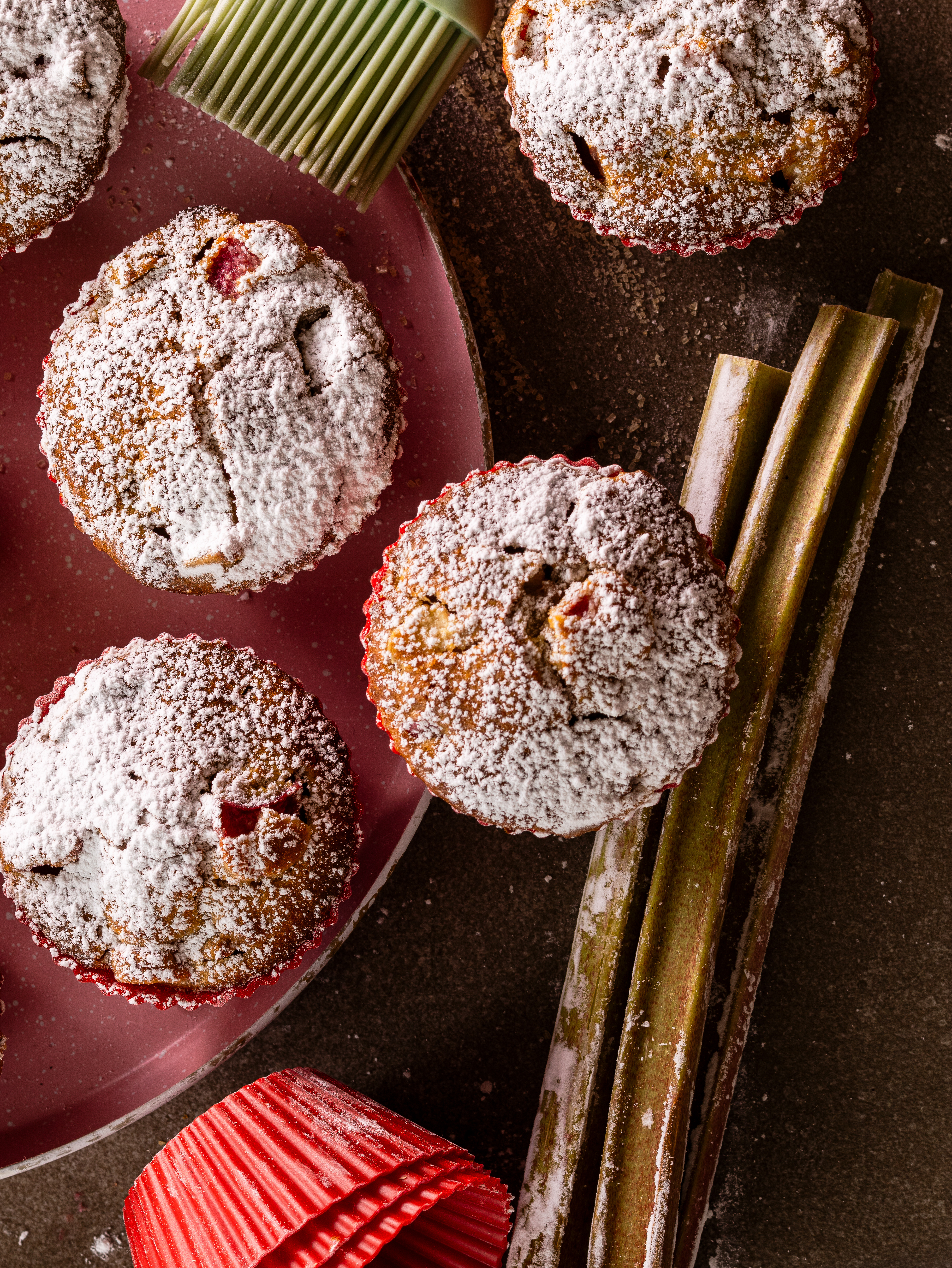 Moody overhead food photograph of rhubarb muffins arranged with fresh rhubarb stalks and red muffin liners.