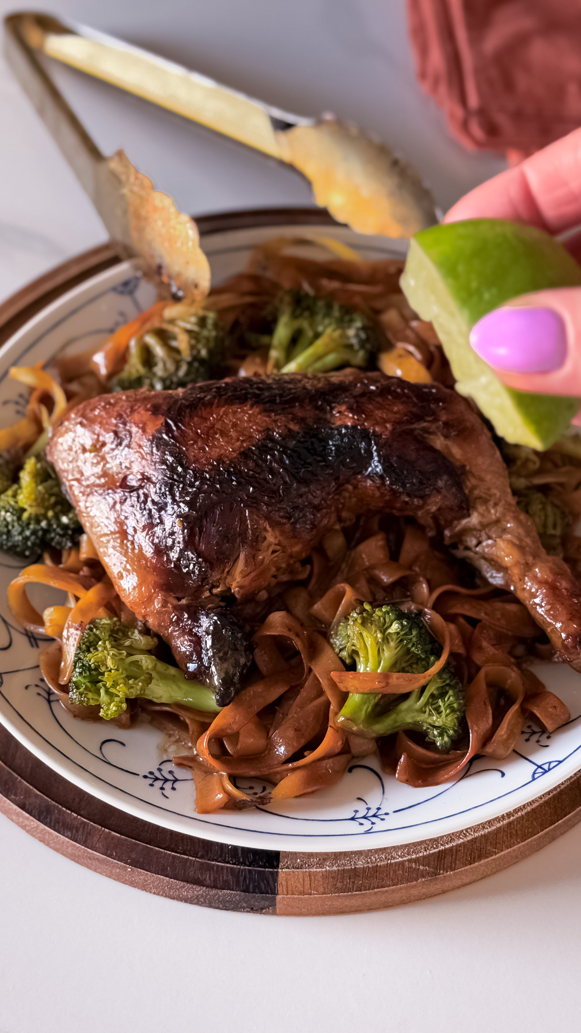 Dark grilled skin chicken leg on a pillow of brown teriyaki tagliatelle and broccoli. A lime halve and large golden tongs.