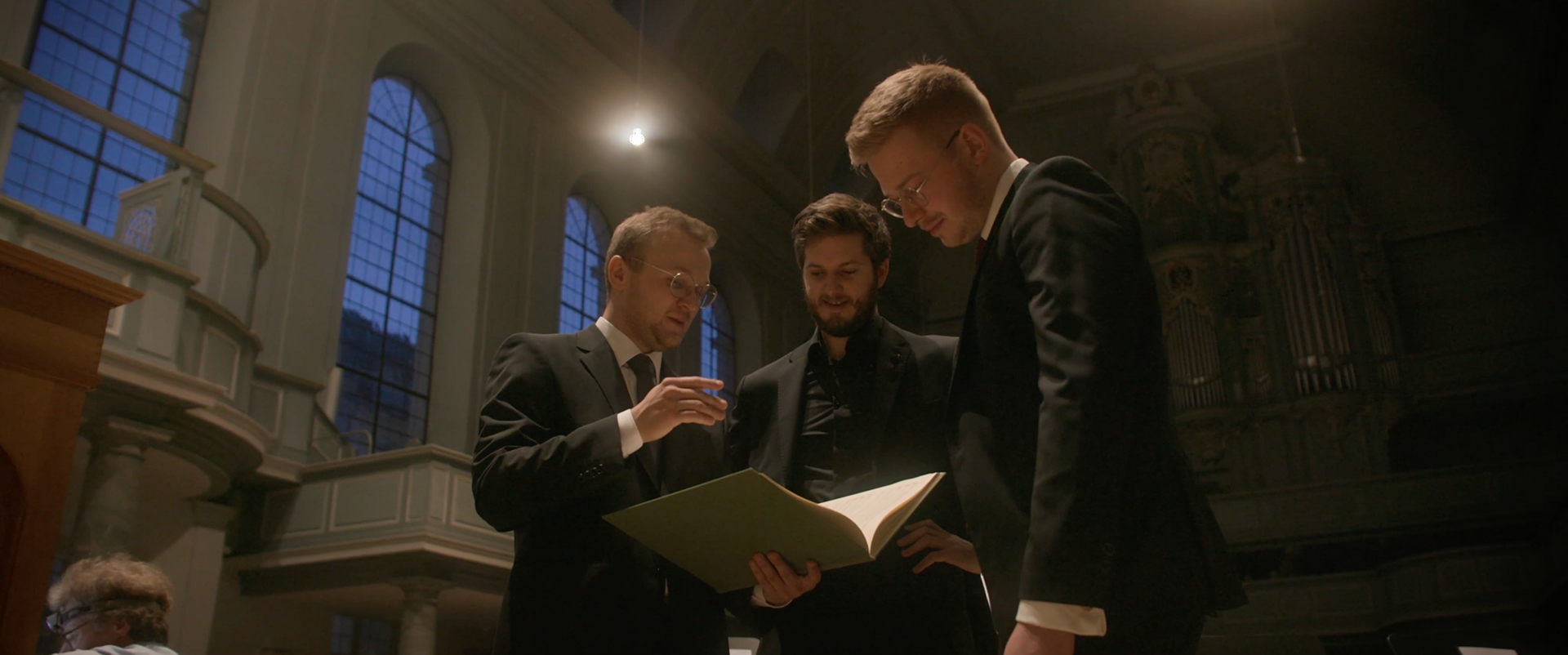 Dirigent Justus Merkel, Alexander Gergelyfi und Manager Philipp Cuno-Friess mit einem Buch in der Kirche St. Gumbertus in Ansbach, Mittelfranken