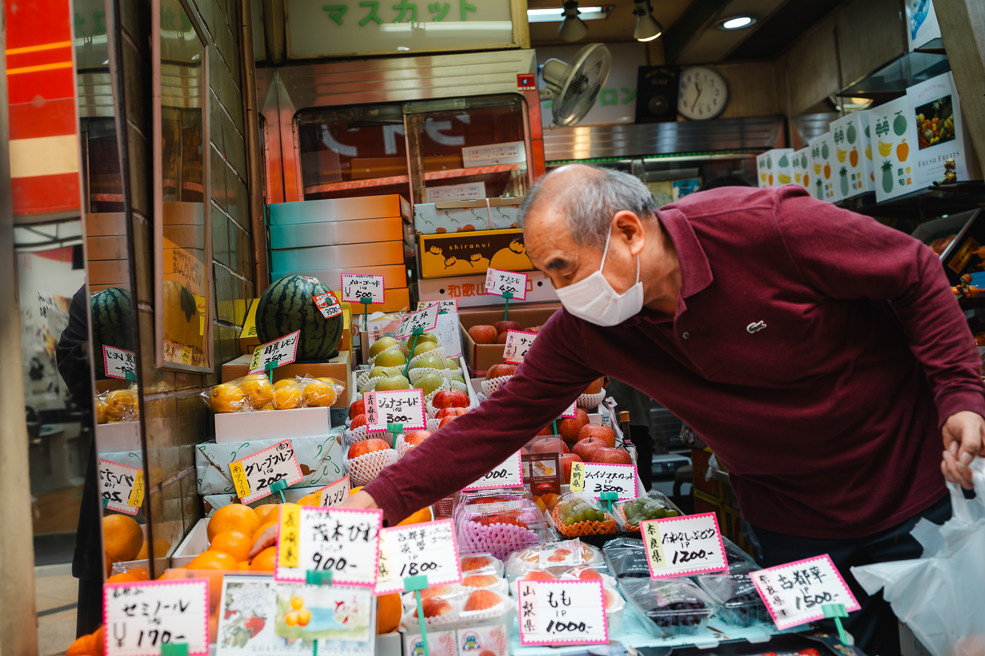 Photographie documentaire par Sabri Kaced montrant un commerçant japonais masqué choisissant des fruits sur un étal coloré dans un marché traditionnel.
