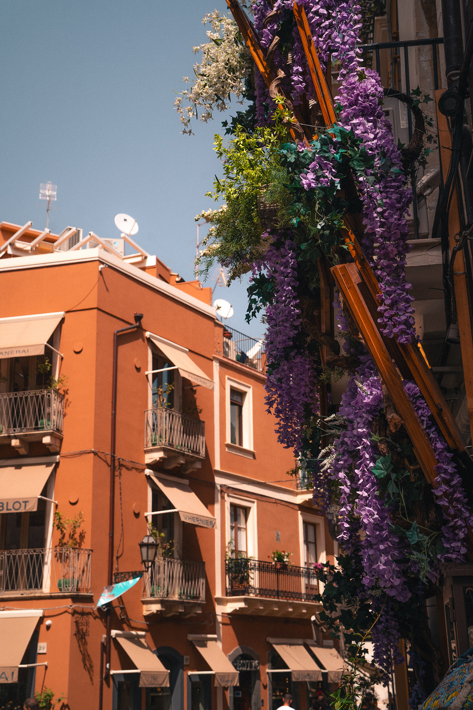 Façade d'un bâtiment orange à Taormine ornée de glycines violettes et de stores, par Sabri Kaced.