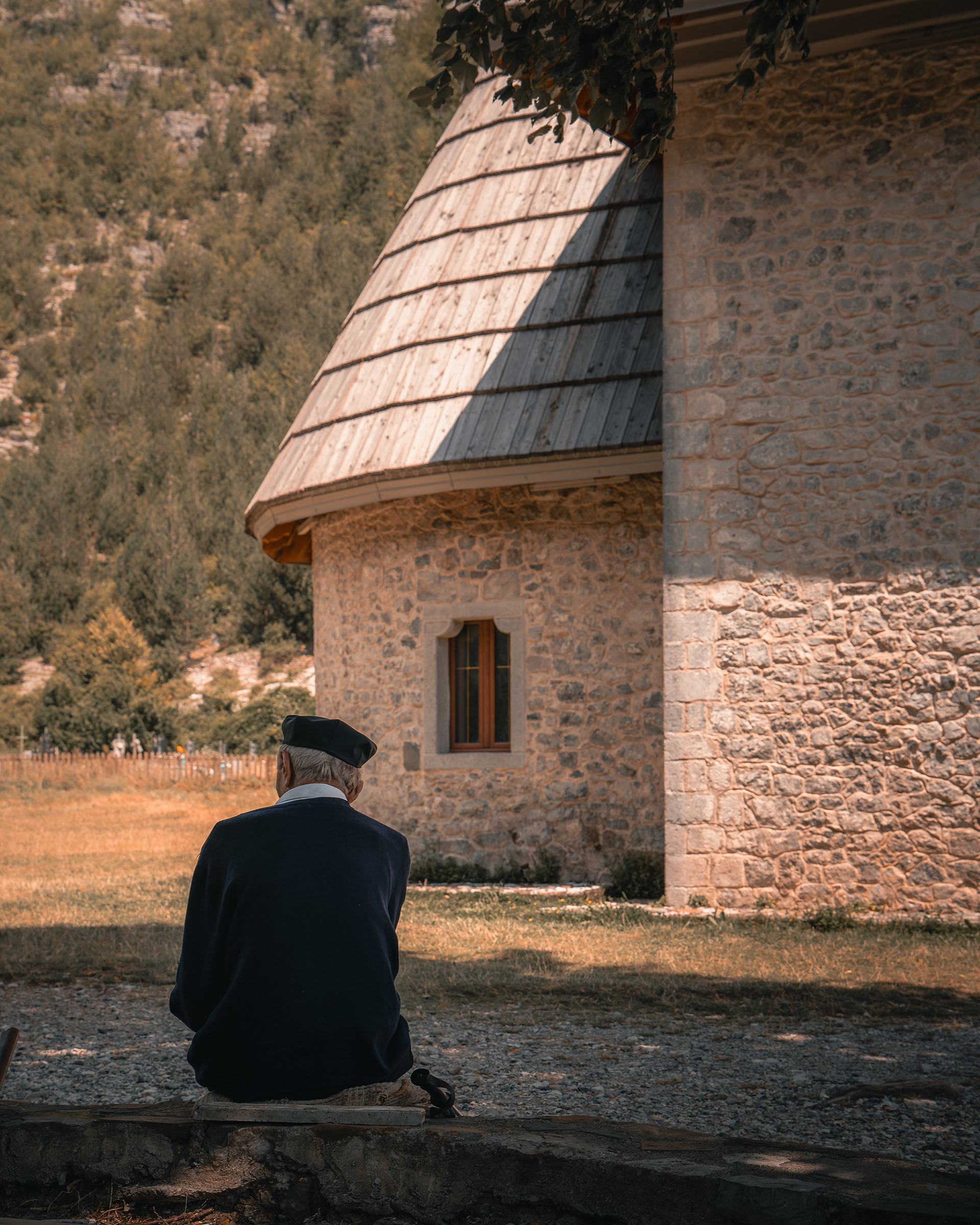Homme âgé portant un béret assis de dos face à une tour en pierre traditionnelle (Kulla) dans le village de Theth, illustrant la culture albanaise. Photo : Sabri Kaced.